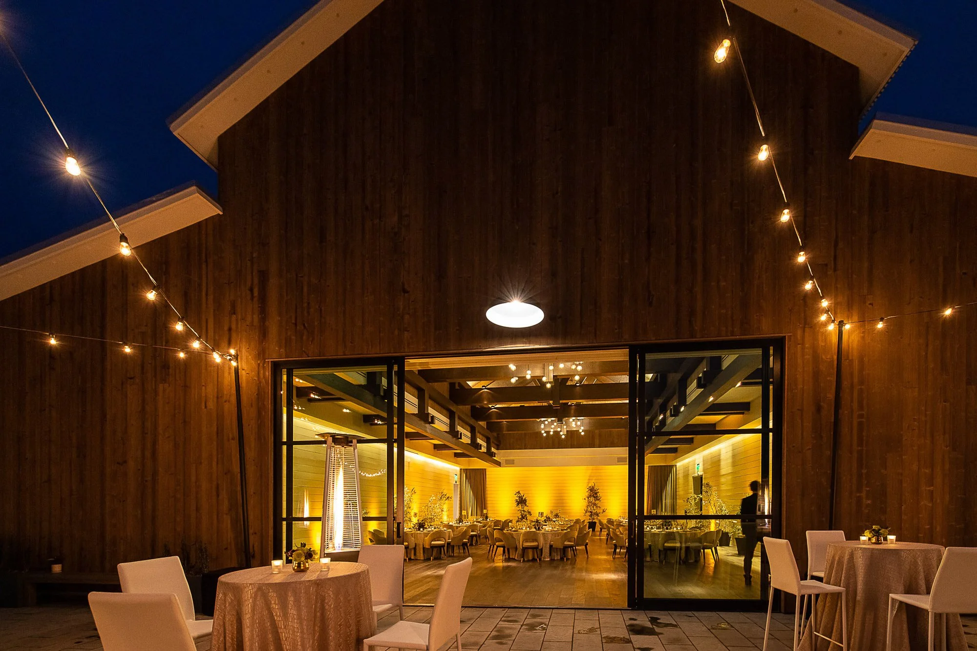 Indoor dining area with round tables covered with beige tablecloths, white chairs, and warm lighting, viewed through an open sliding door at night
