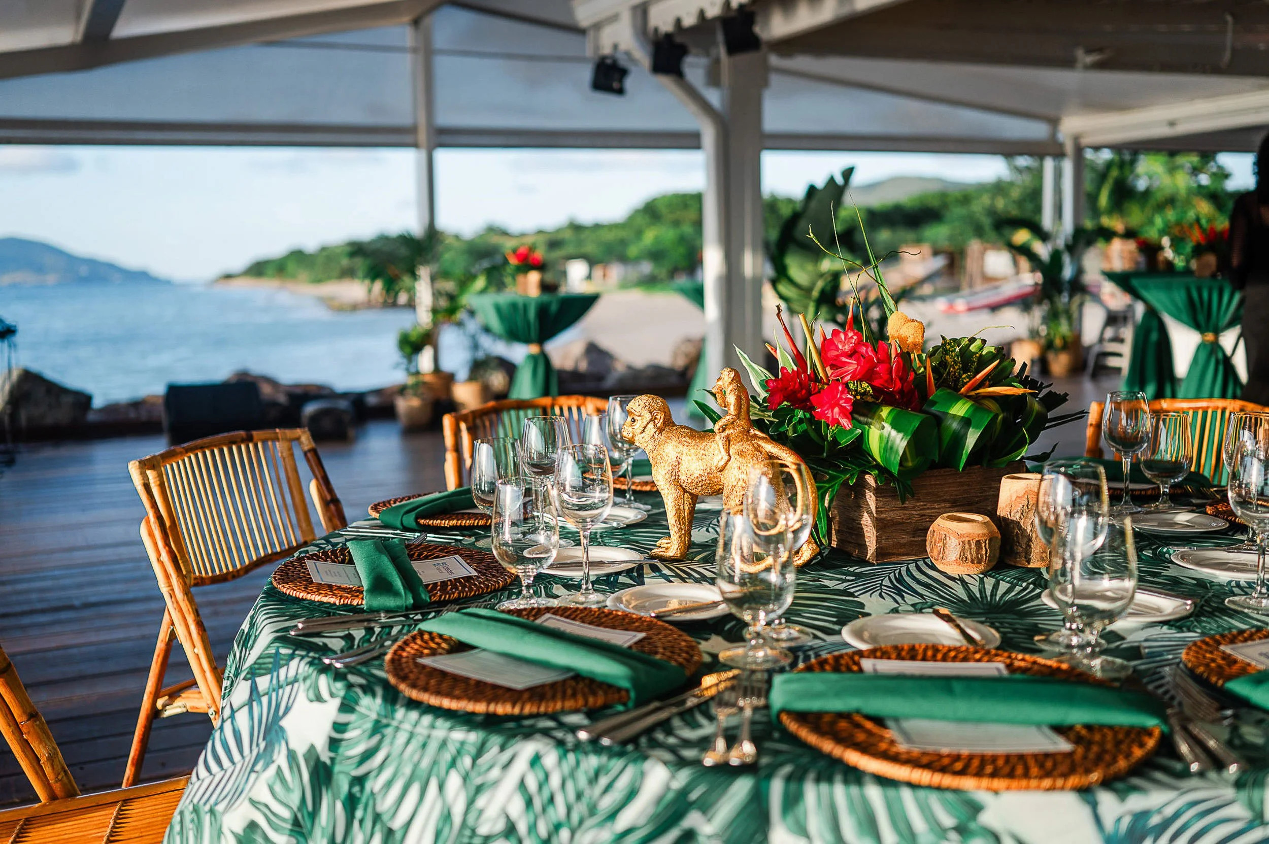 A round table set for a meal with floral arrangements, gold elephant figurines, napkins, glassware, and plates, on an outdoor deck overlooking the water with a distant shoreline.
