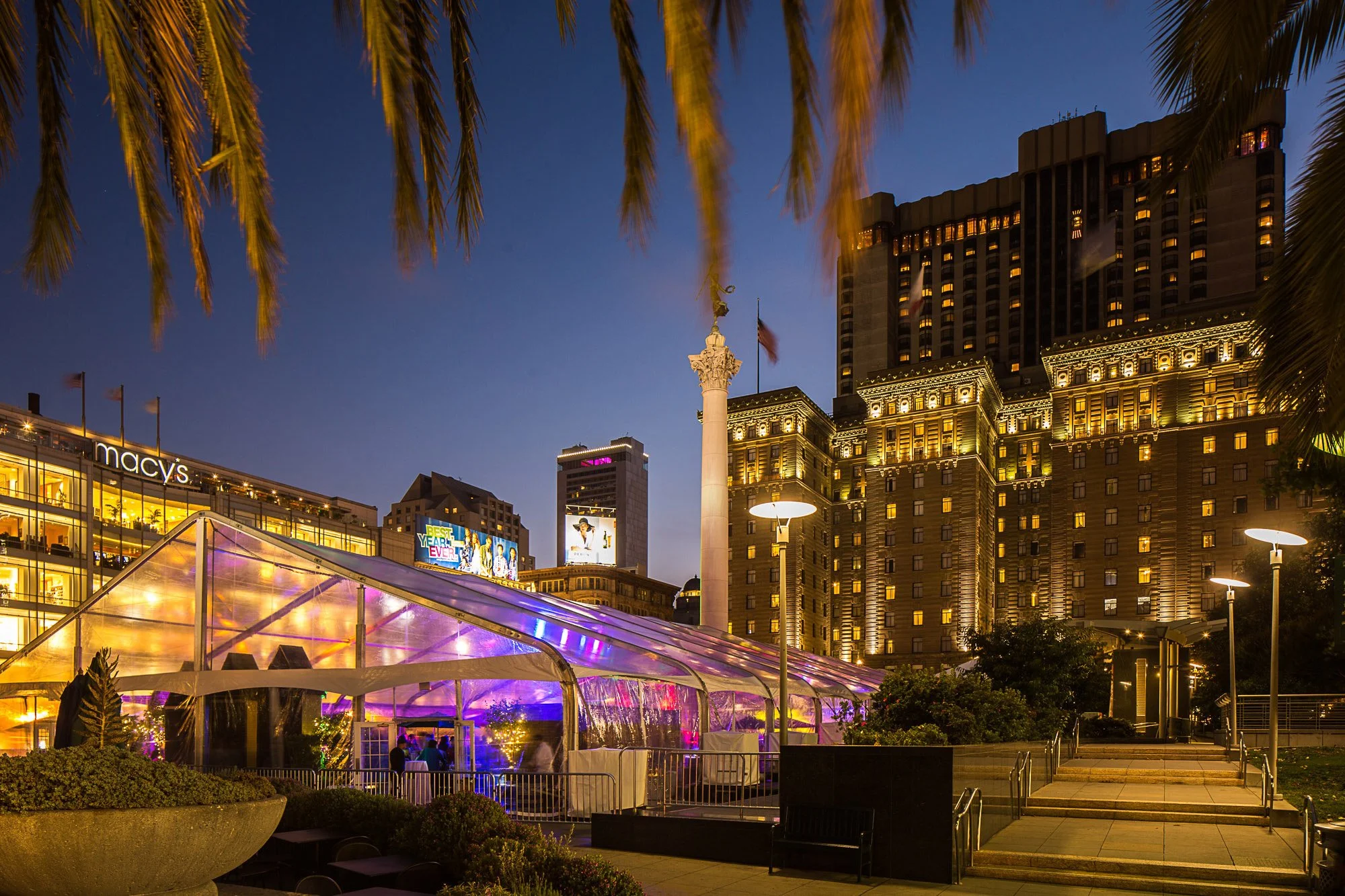 Night view of Las Vegas city blocks with illuminated buildings, palm fronds at the top, a Macy's store, and a transparent purple-lit tent in the foreground.