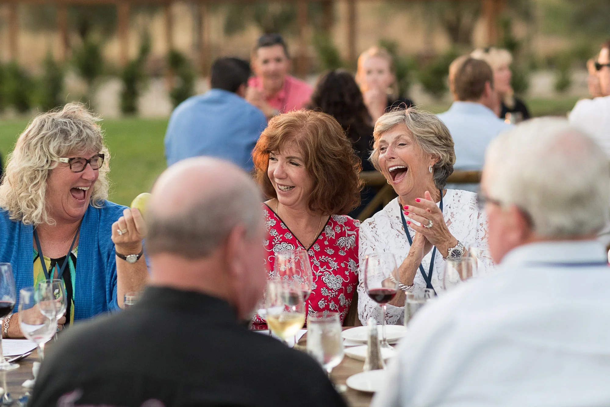 People gathered around a long table at an outdoor event, smiling and laughing, with glasses of wine and plates of food in front of them, enjoying a lively conversation.