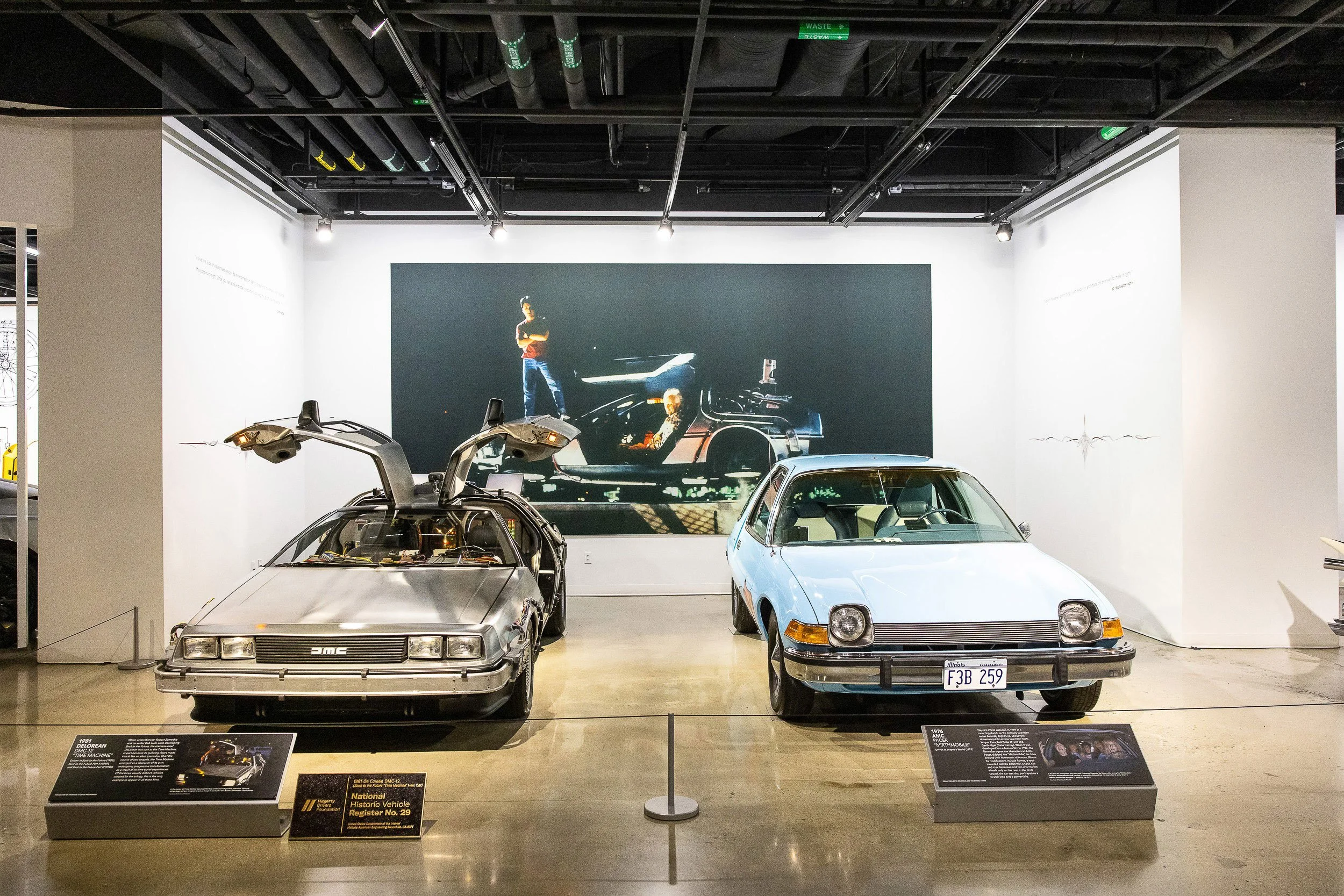 The image shows a car museum exhibit featuring two vintage DeLorean time machines from the movie "Back to the Future" and other classic cars. There are informational plaques in front of the cars and a large picture of the DeLorean with characters fro