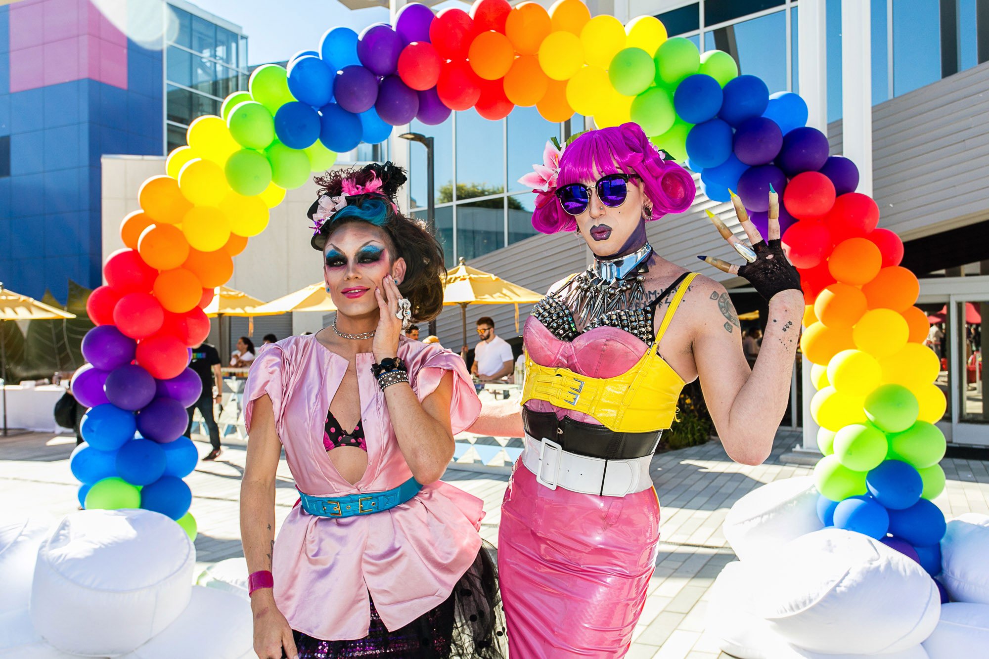 Two individuals dressed in colorful, vibrant costumes standing under a rainbow-colored balloon arch at an outdoor event.