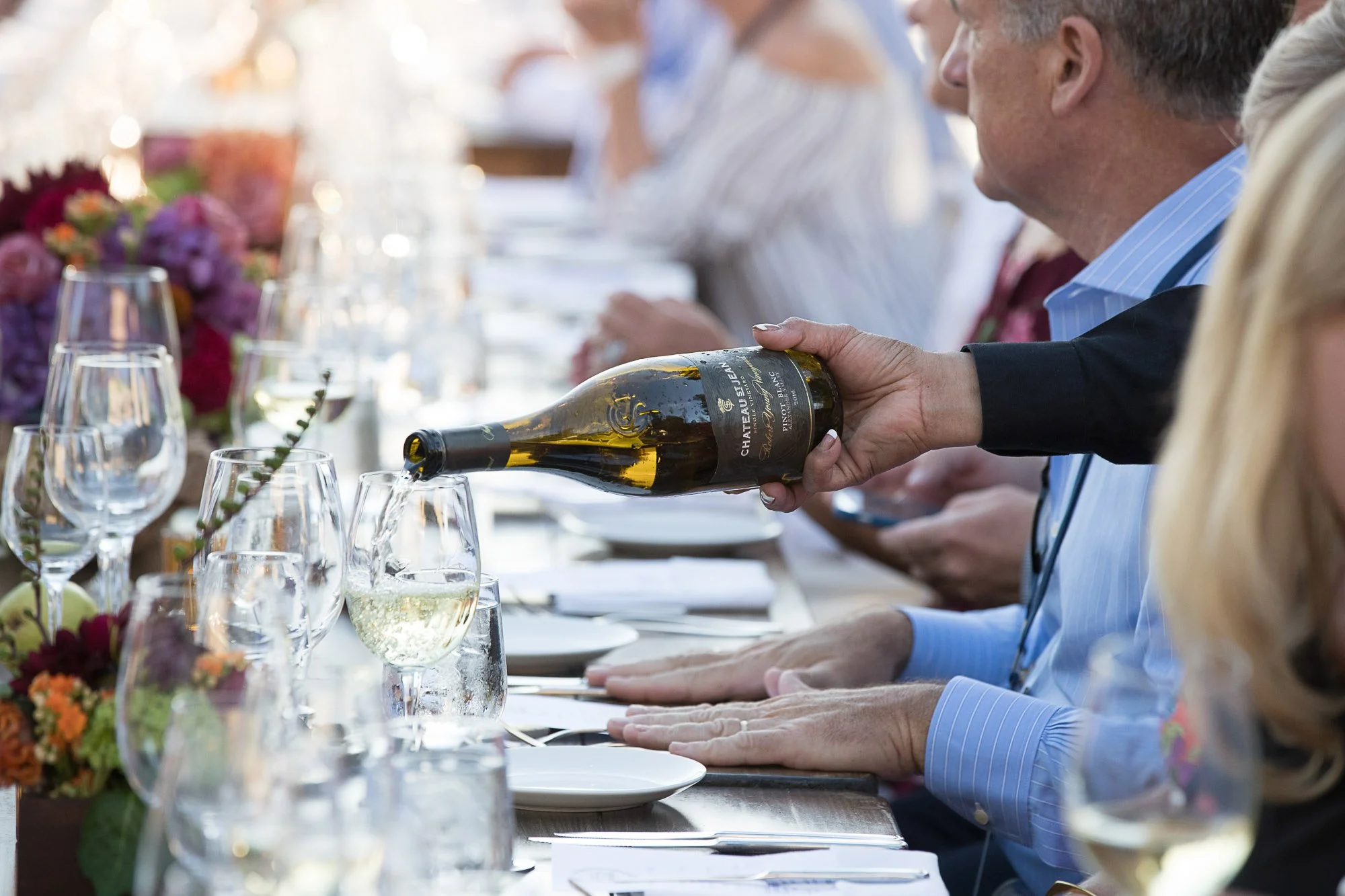 People sitting at a dining table during a meal, with a person pouring white wine into a glass, in an outdoor setting decorated with flowers.