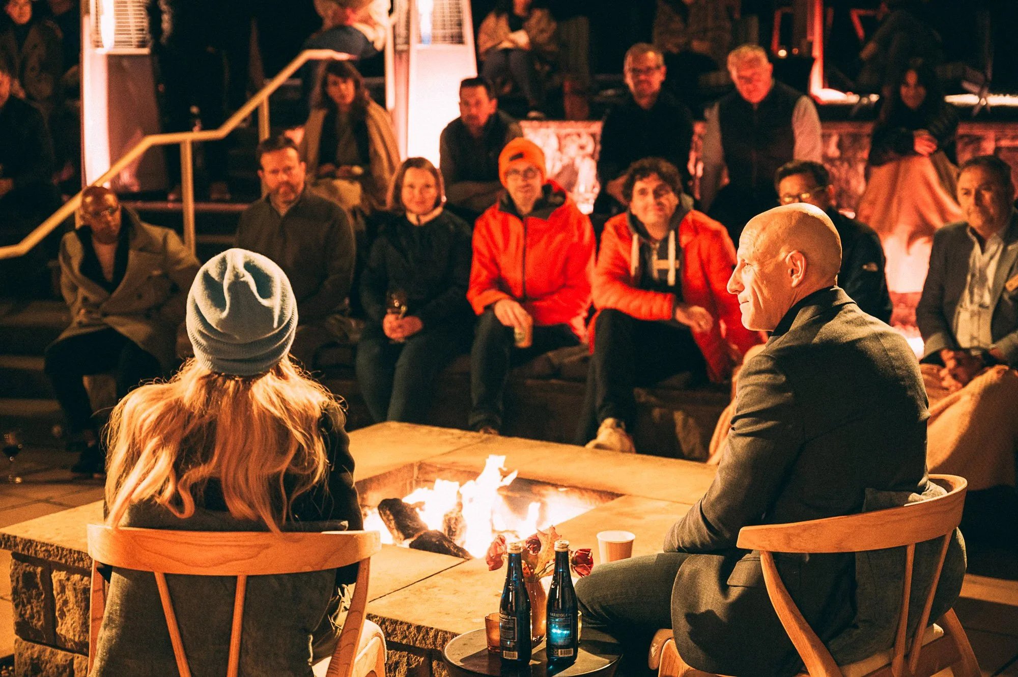 A group of people sitting around a fire table outdoors at night, socializing and enjoying drinks.