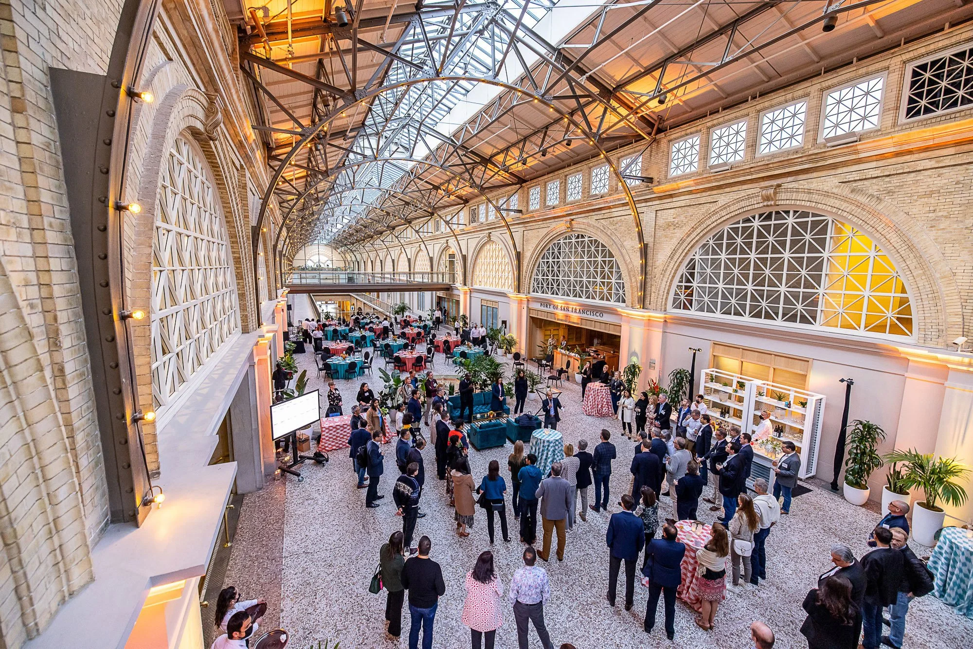 An indoor event at the Port of San Francisco in a spacious historic building with high arched glass ceiling and brick walls, where people are gathered around tables and socializing.