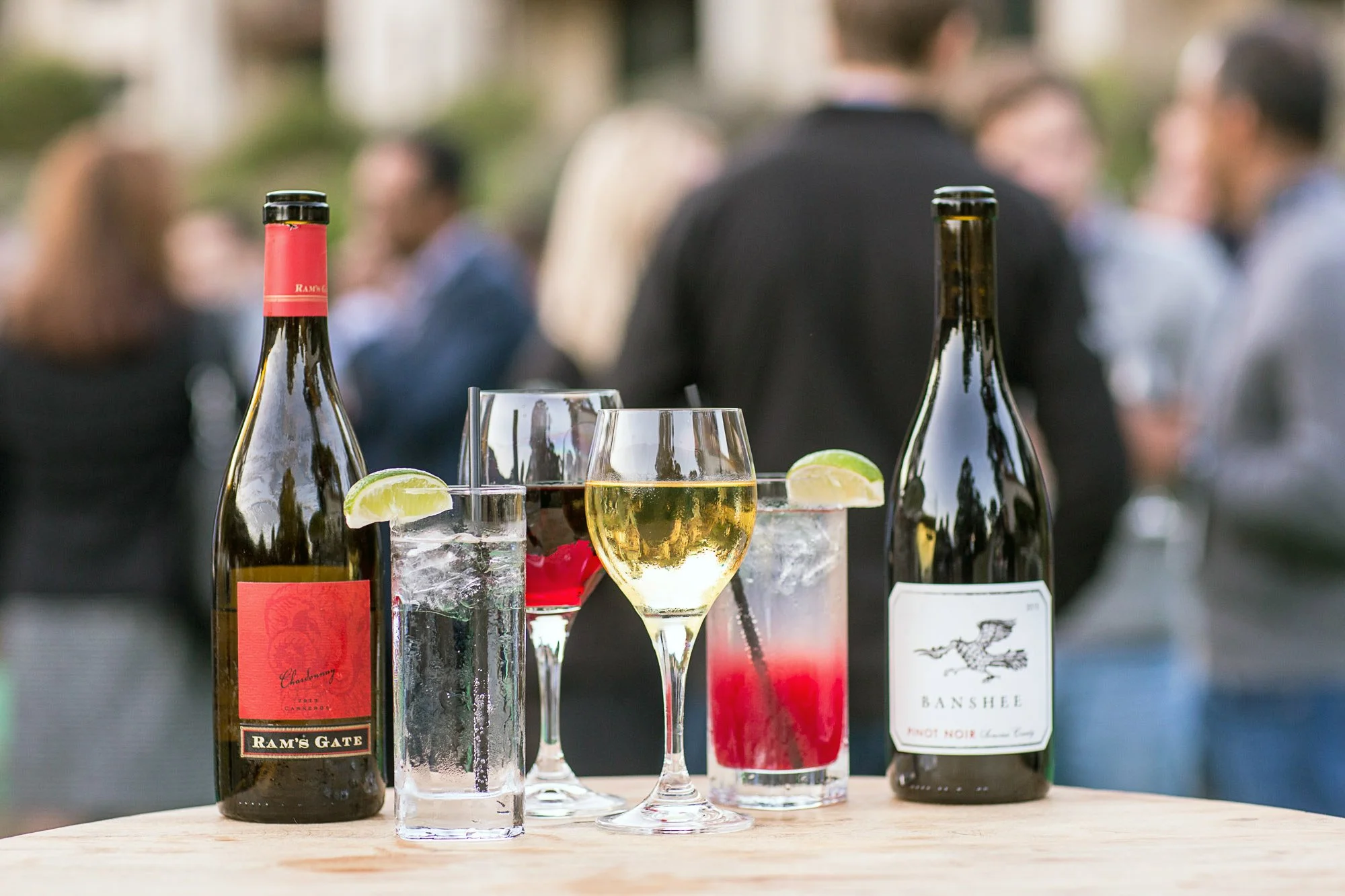 Three bottles of wine and several glasses of drinks with lime wedges on a wooden table at an outdoor gathering. Background shows blurred people socializing.