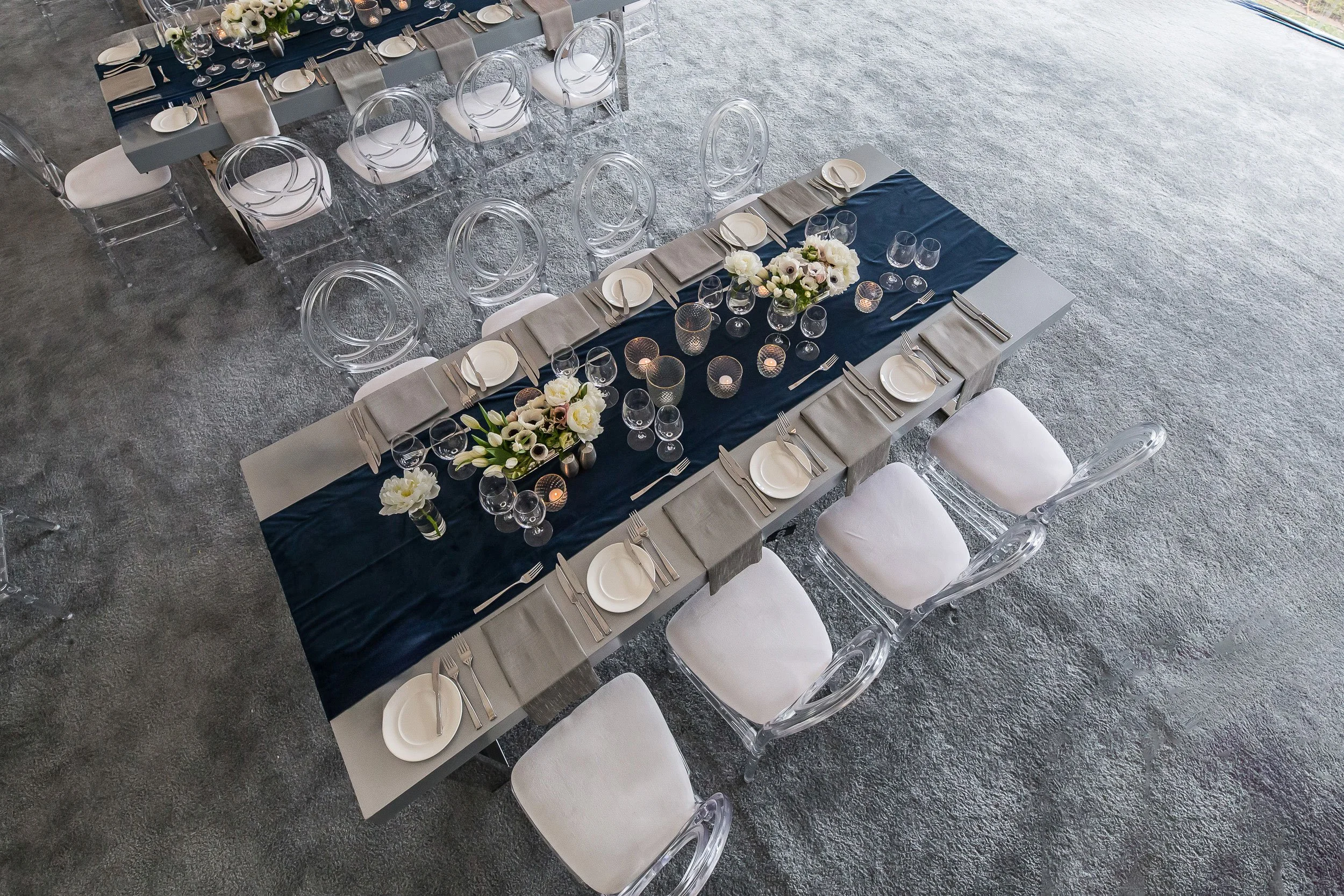 Overhead view of a formal dining table decorated with a dark blue runner, floral centerpieces, candles, and place settings including plates, silverware, glasses, and folded napkins. Transparent chairs with white cushions are arranged around the table
