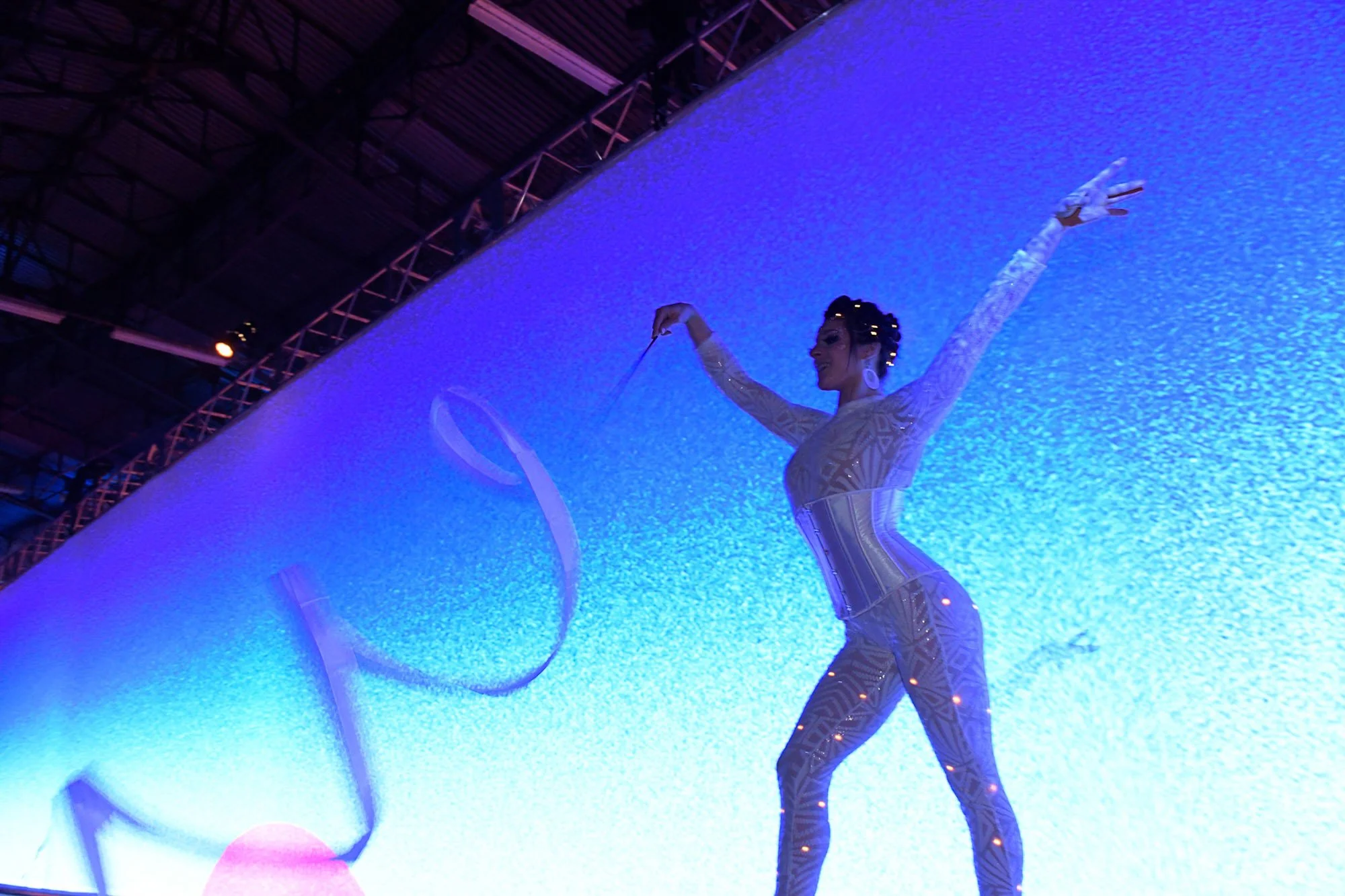 A woman performing a hoop dance on a stage with blue lighting, wearing a patterned bodysuit, with her arms raised and a smiling expression.