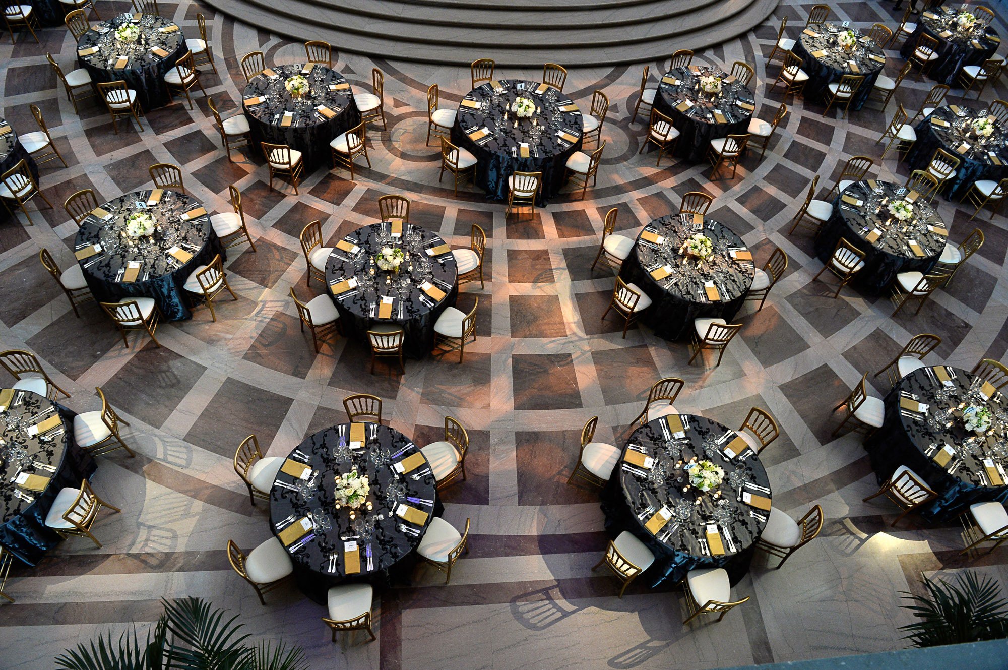 Multiple round banquet tables with black and gold chairs, decorated with floral centerpieces and set with glassware, silverware, and yellow napkins, arranged in a large, elegant banquet hall.