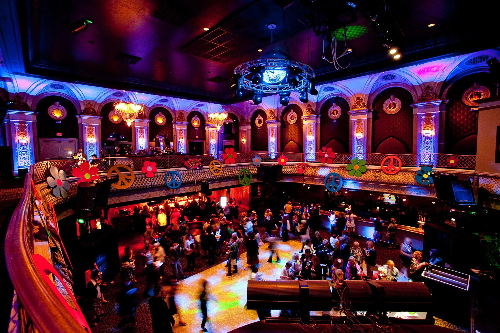 A lively indoor nightclub or event space with colorful lighting, ornate gold and white architectural details, and a large crowd of people dancing and socializing on the main floor. There are decorative flower and peace sign cutouts hanging along the 