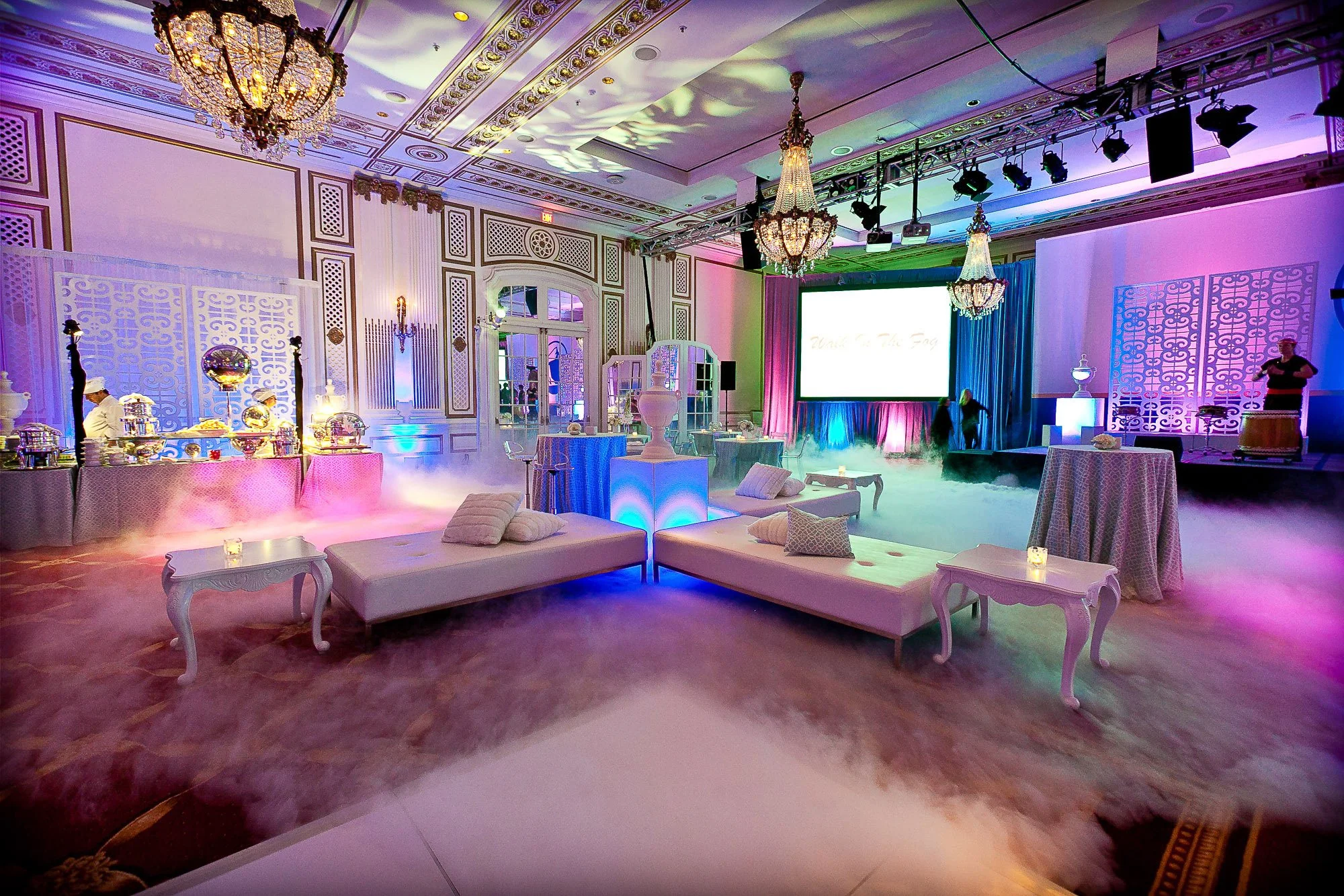 A large ornate banquet hall decorated with chandeliers, colorful lighting, and a stage with a large screen, set up for an event.