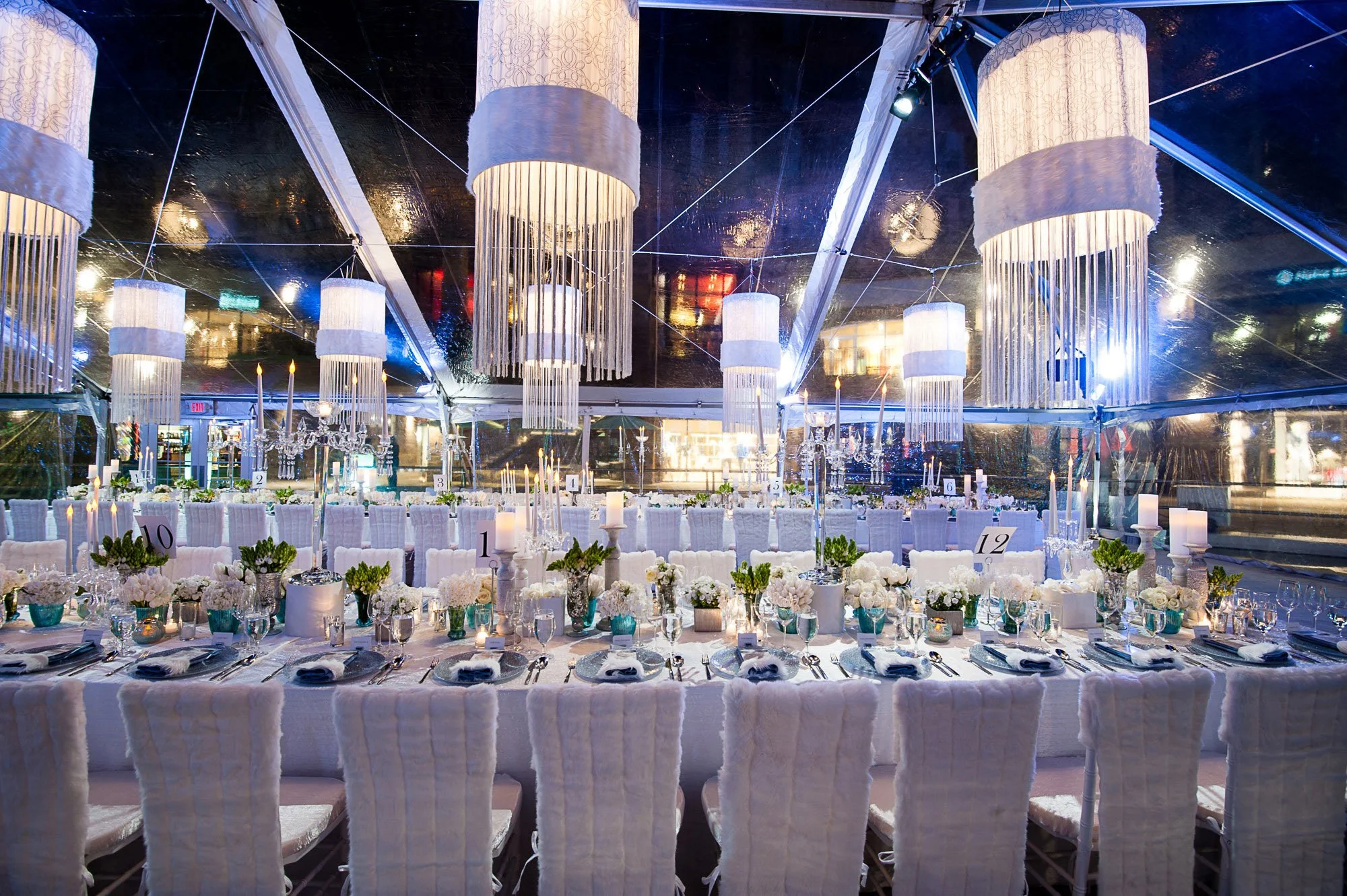 Elegant banquet setup inside a clear tent with white chairs, floral centerpieces, and candlelit table, illuminated by large hanging chandelier-style lights.