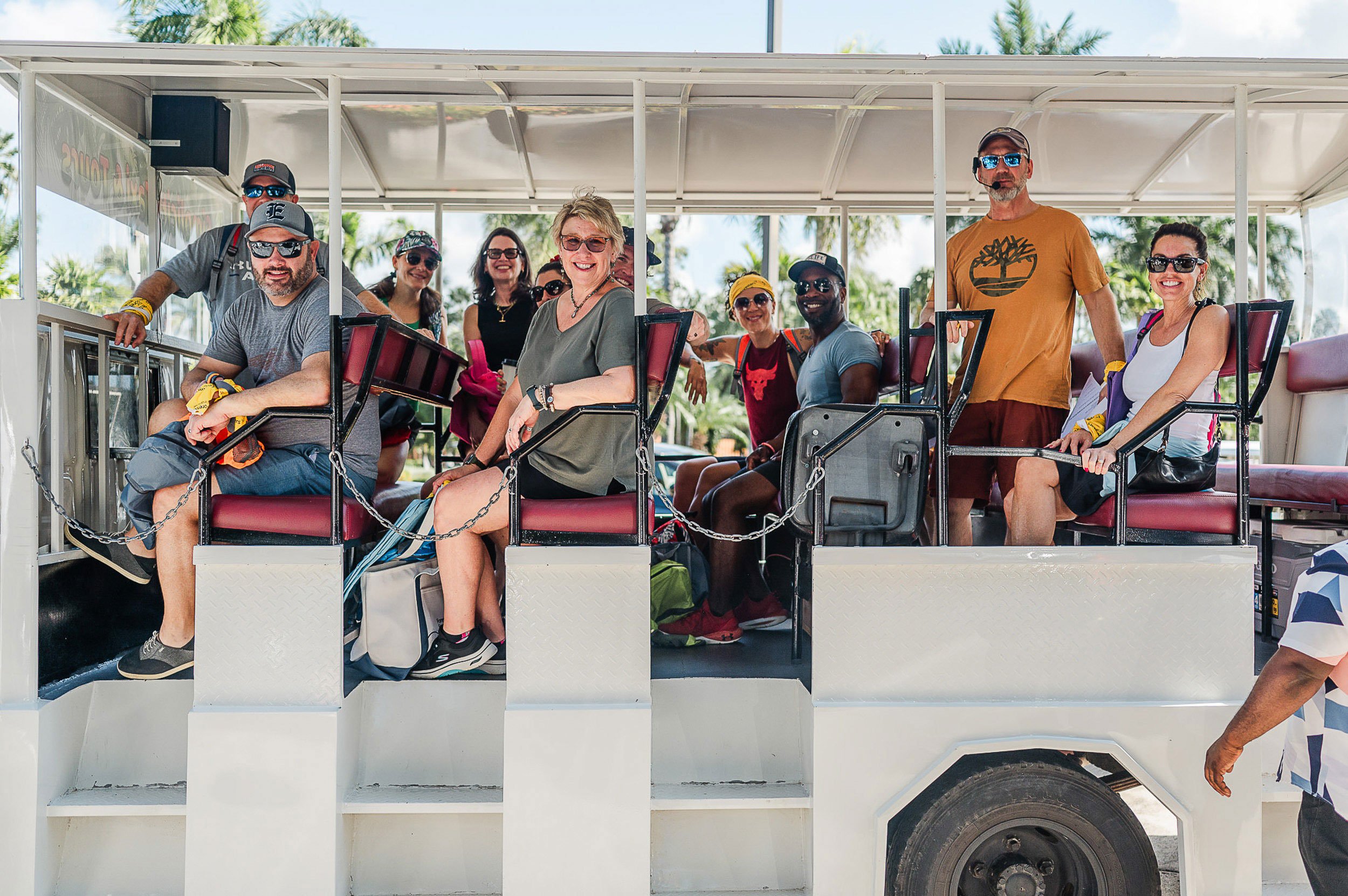 A group of people sitting and standing on a white open-air trolley or tram, smiling at the camera, with palm trees in the background and clear blue sky.