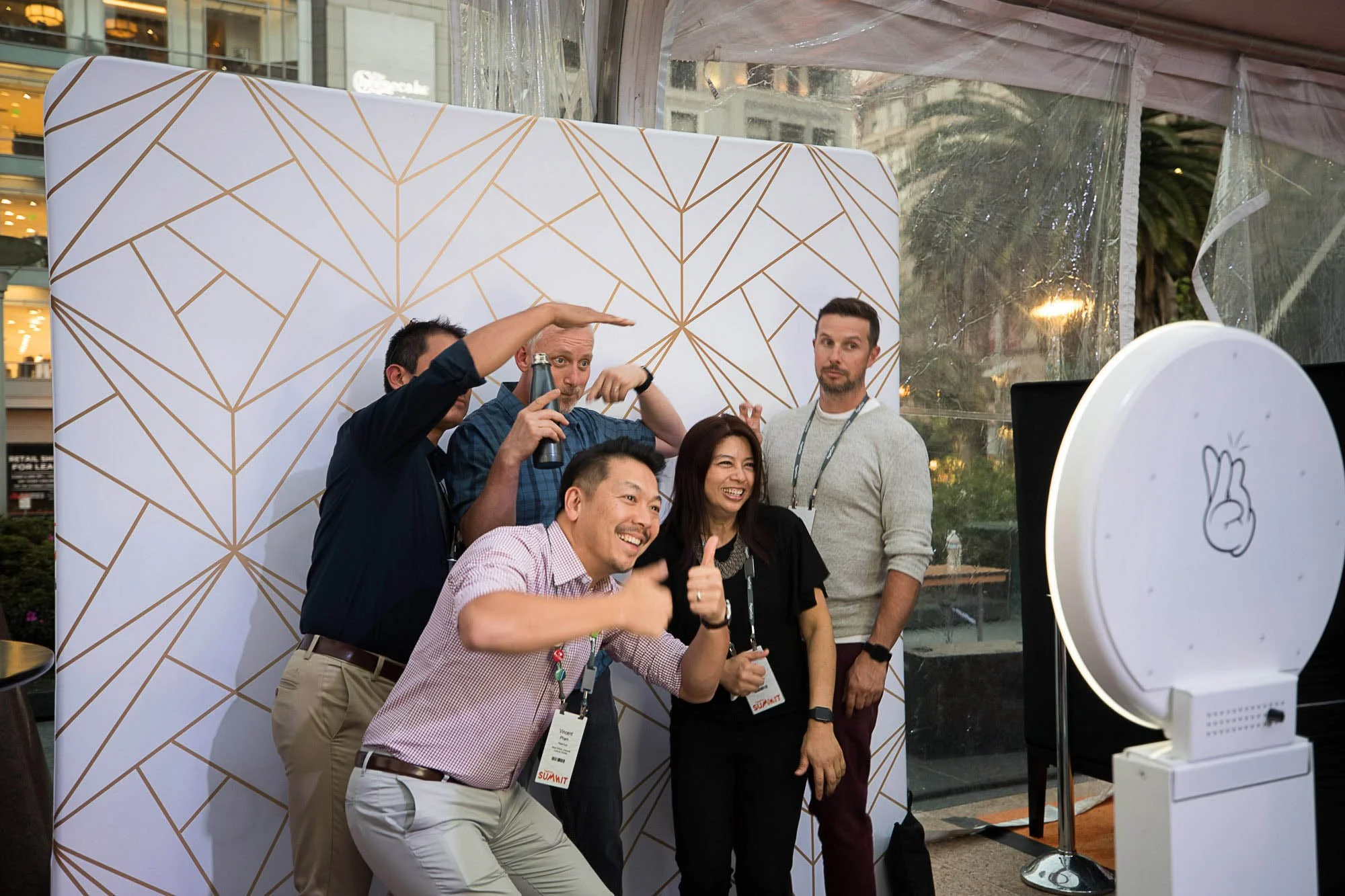 Group of people posing for a photo at a professional event or conference, standing in front of a decorative backdrop, with one person giving a thumbs-up and others smiling and holding photo props.