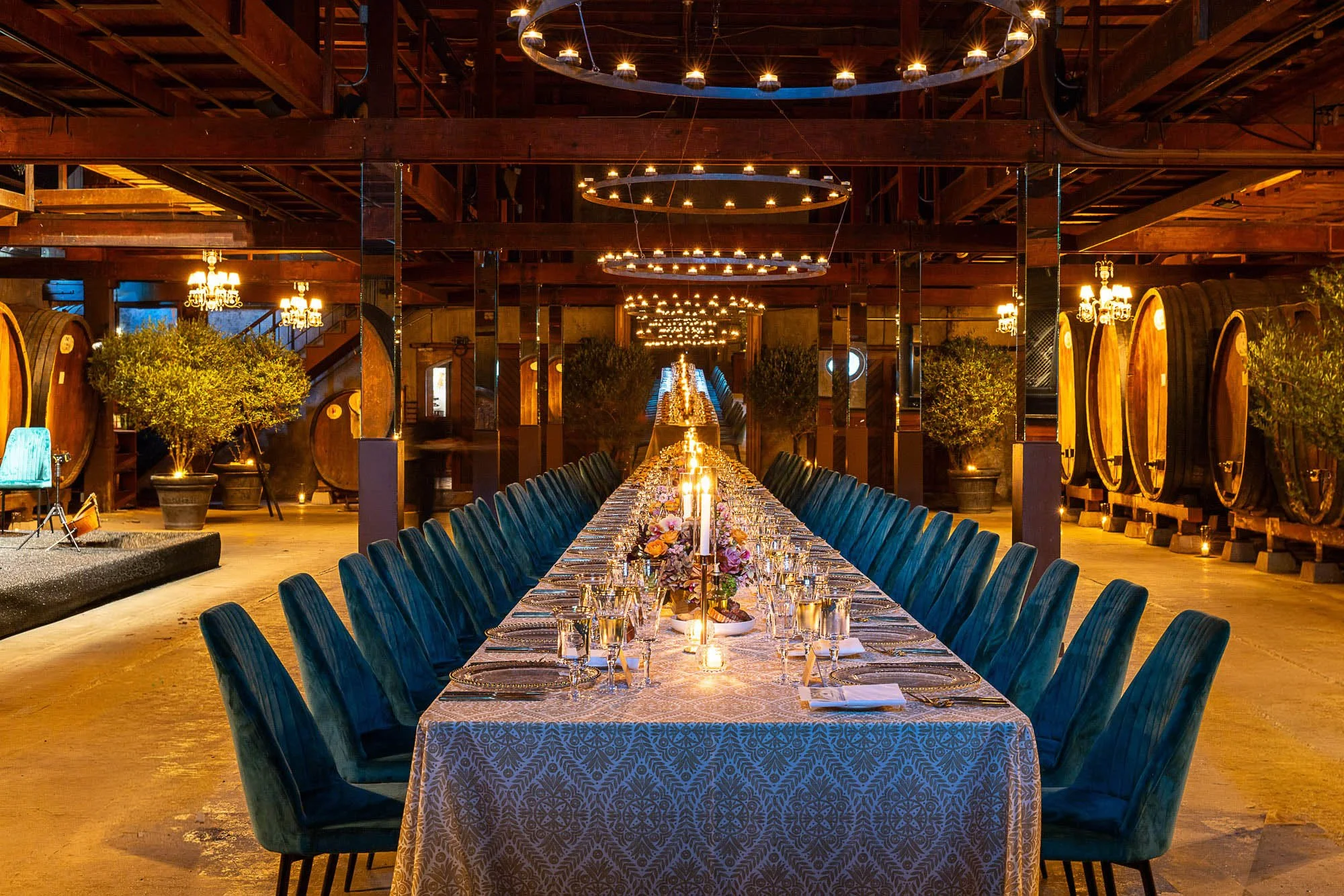 A long banquet table set with floral arrangements and candles in a rustic, elegant dining space with wooden barrels, greenery, and chandeliers hanging from the ceiling.