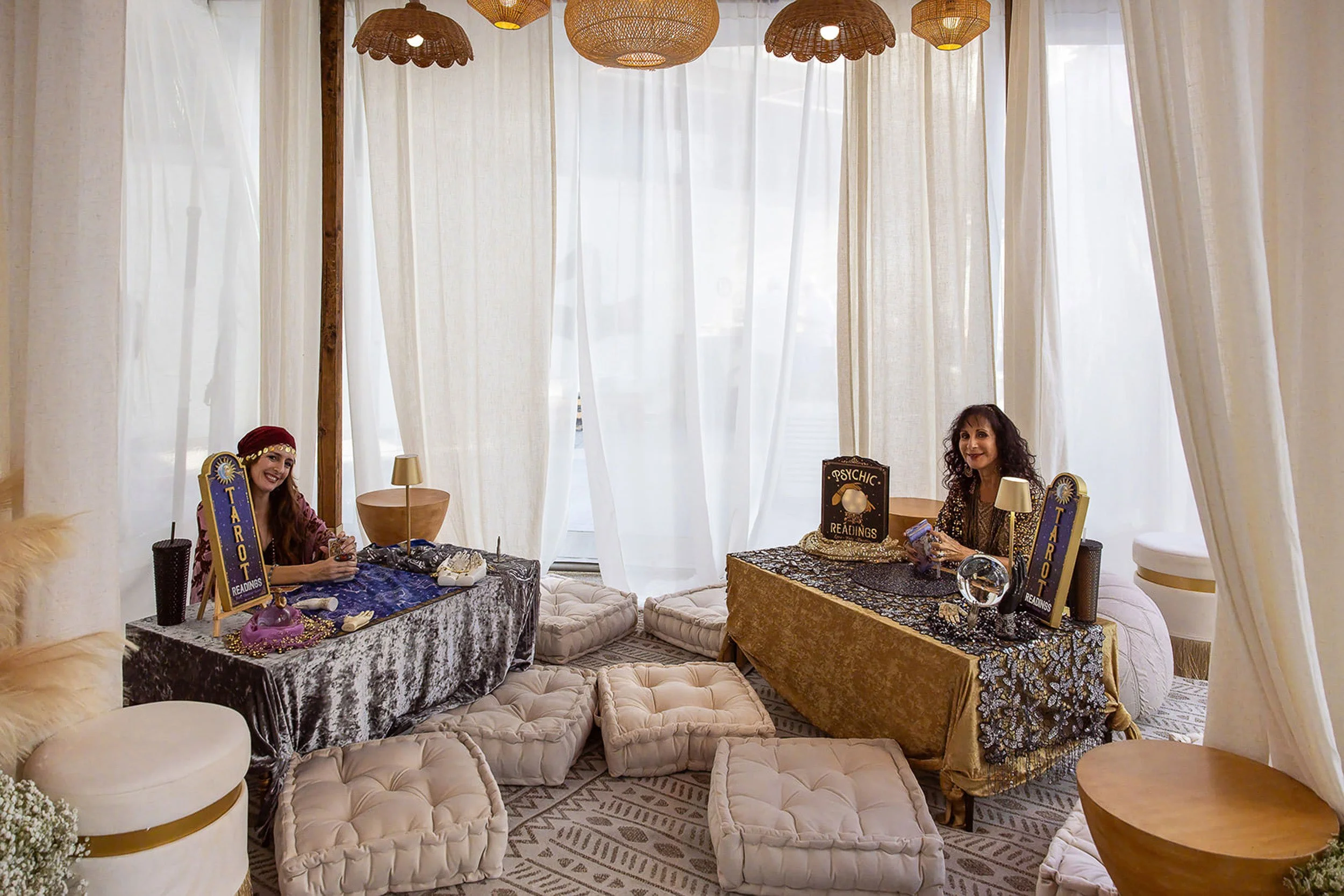 Two women sitting at small tables with tarot reading signs, decorated with crystals, tarot cards, and jewelry, in a cozy room with cream-colored curtains, woven pendant lights, and cushioned seating.