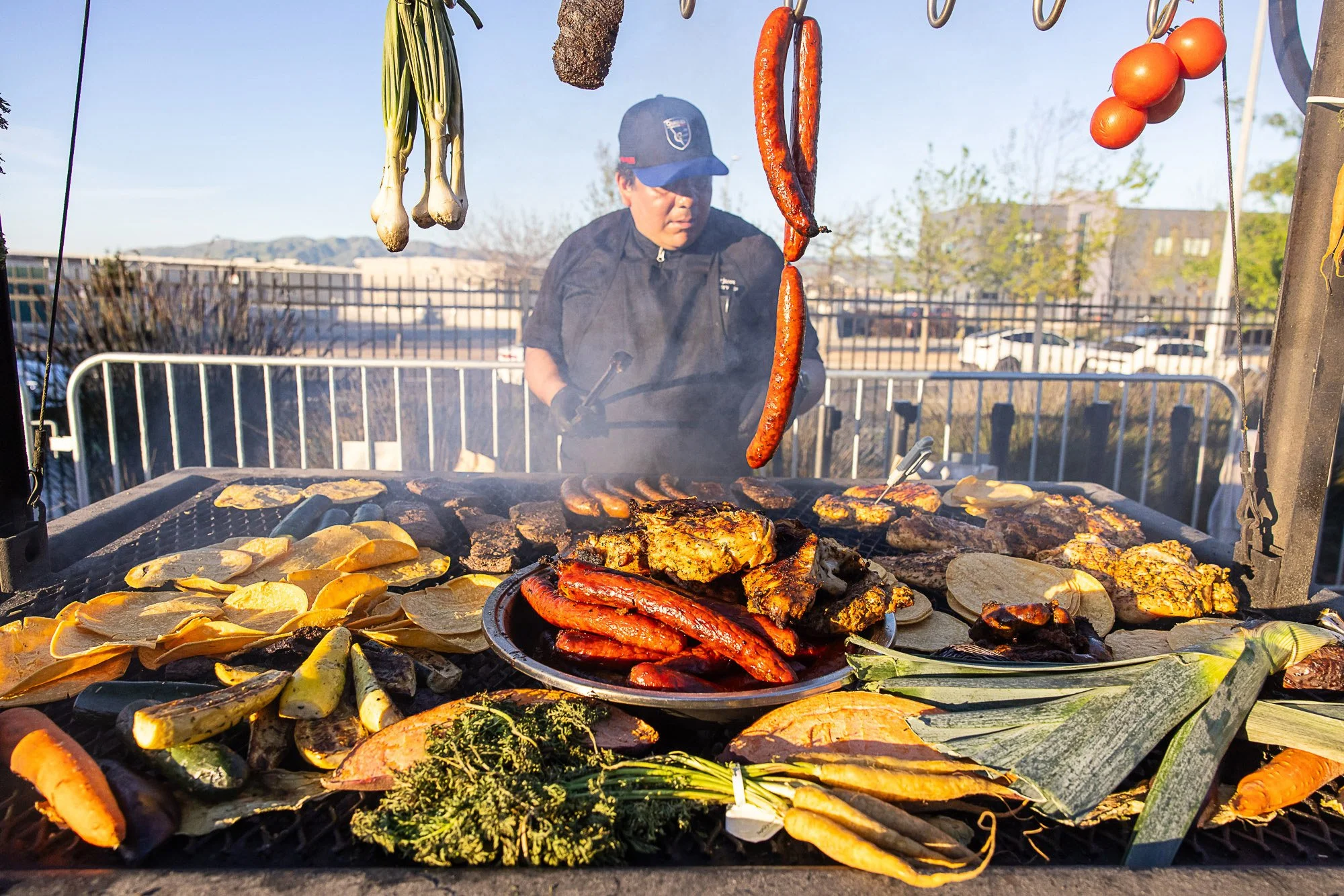 Barbecue grill with sausages, ribs, chicken, sliced vegetables, and flatbreads cooking outdoors on a sunny day, with a man wearing a blue cap and black jacket observing in the background.