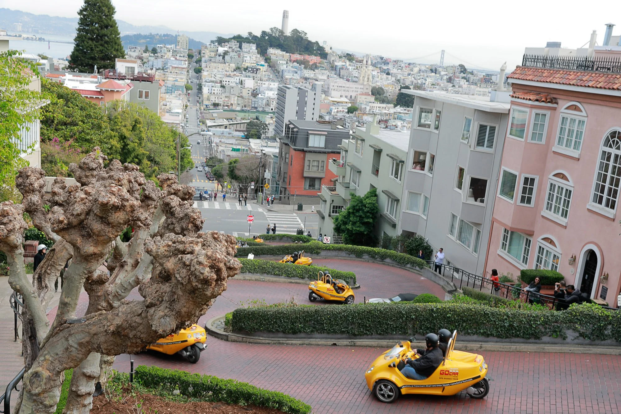 A view of a San Francisco street with colorful buildings, people walking, and yellow GoCar rental vehicles in the foreground, including a person driving one.