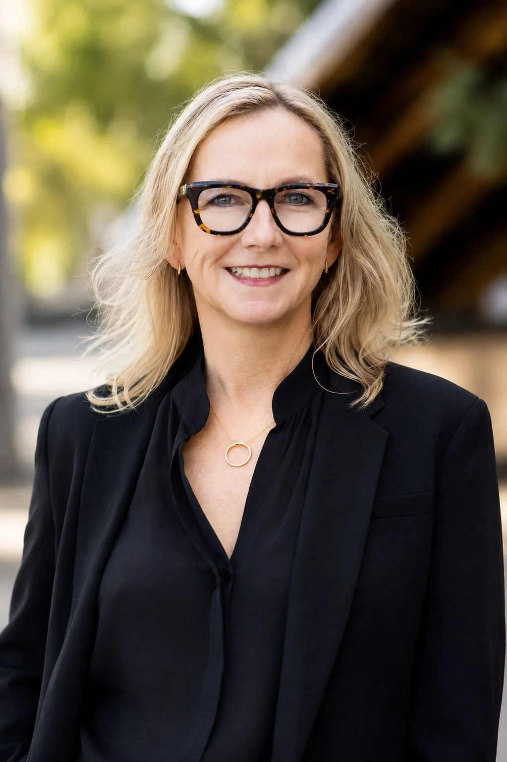 A woman with blonde, wavy hair and glasses smiling outdoors, wearing a black blazer and necklace.