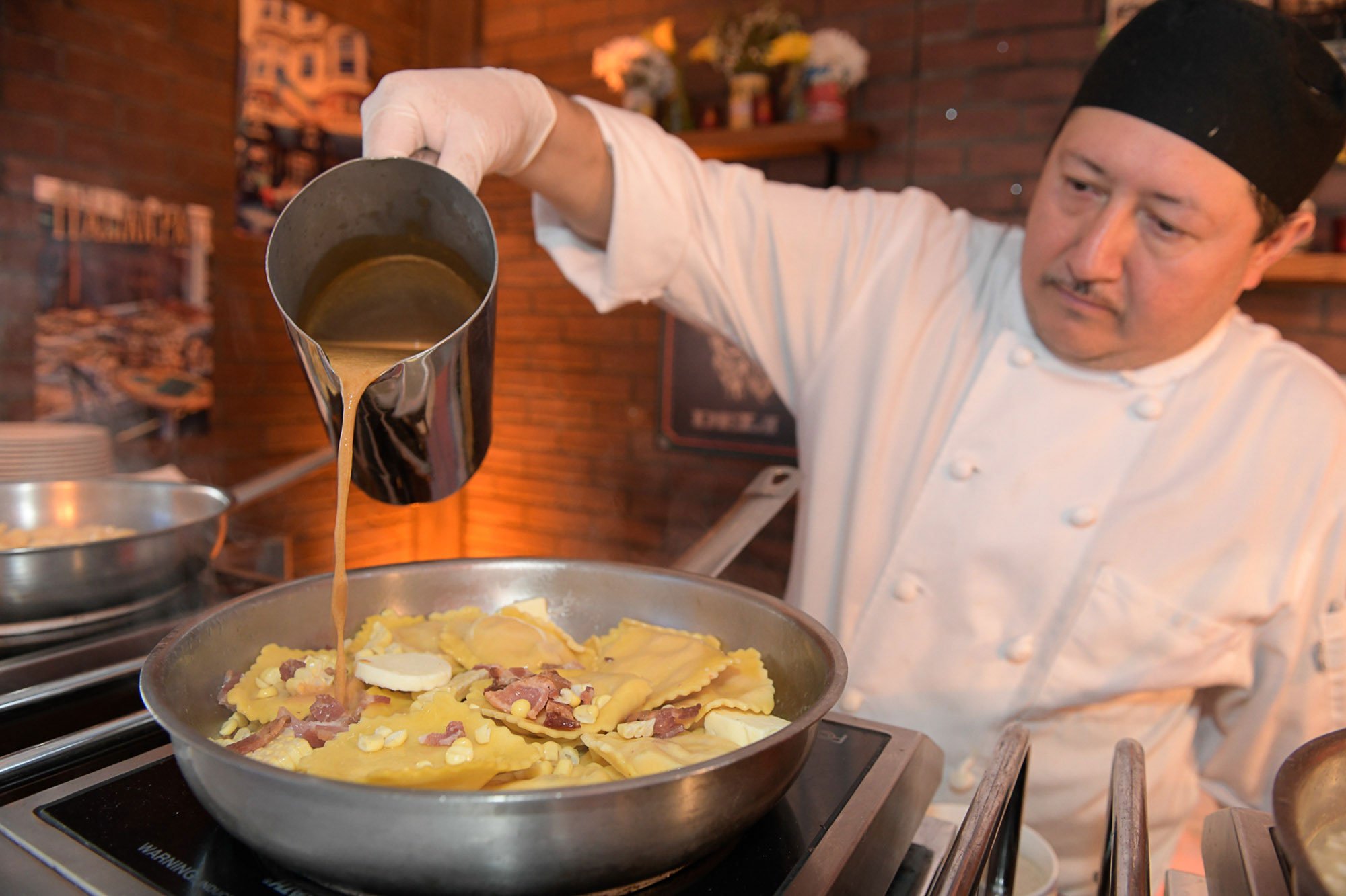 A chef pouring gravy over a dish of fresh pasta topped with cheese, bacon, and eggs in a restaurant kitchen.