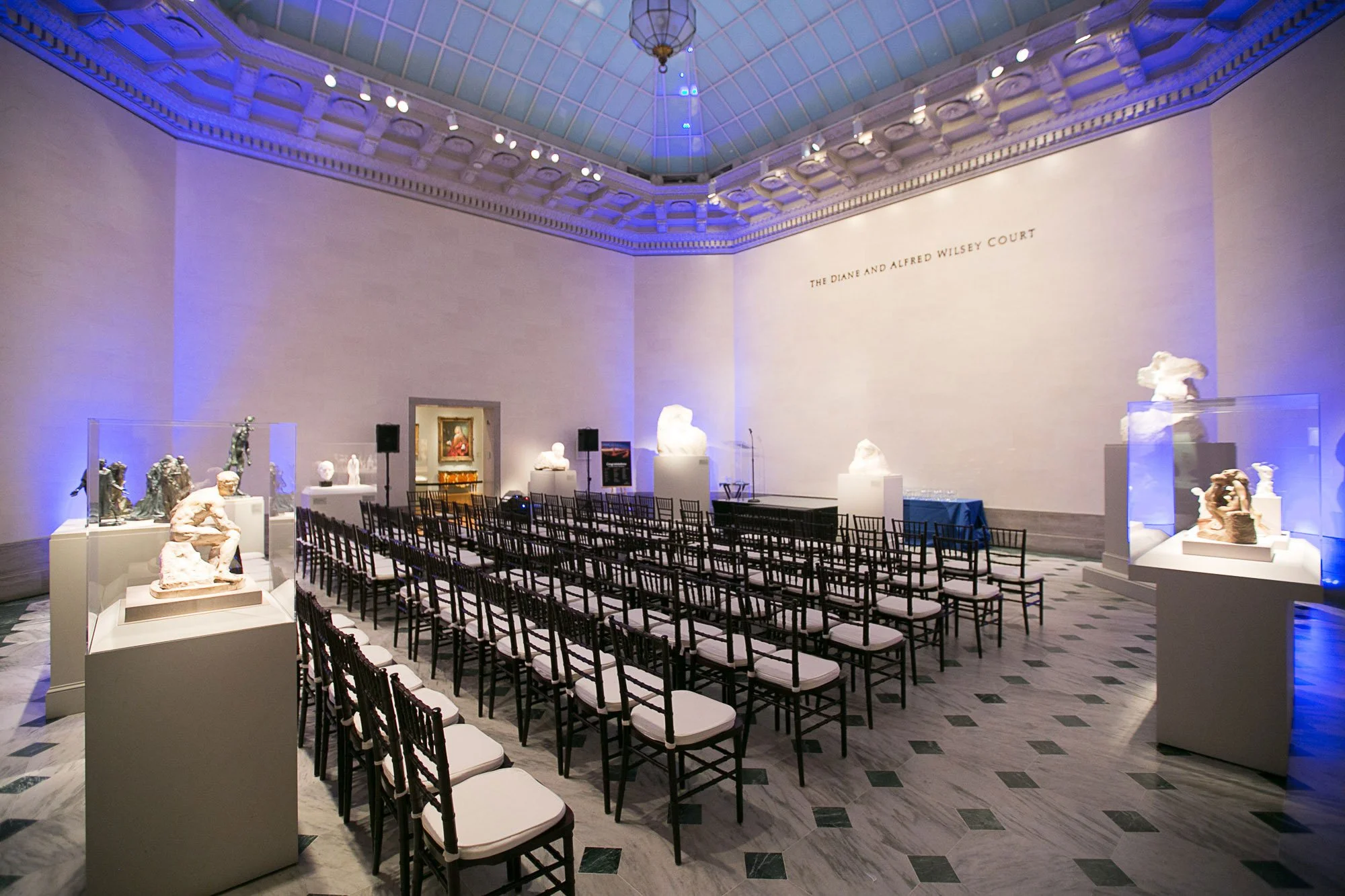 Interior of a museum or art gallery with sculptures displayed on pedestals, rows of chairs set up for an event, and a stage area. The space has high ceilings with decorative moldings and a large inscription reading 'The Diane and Alfred Wilesey Court