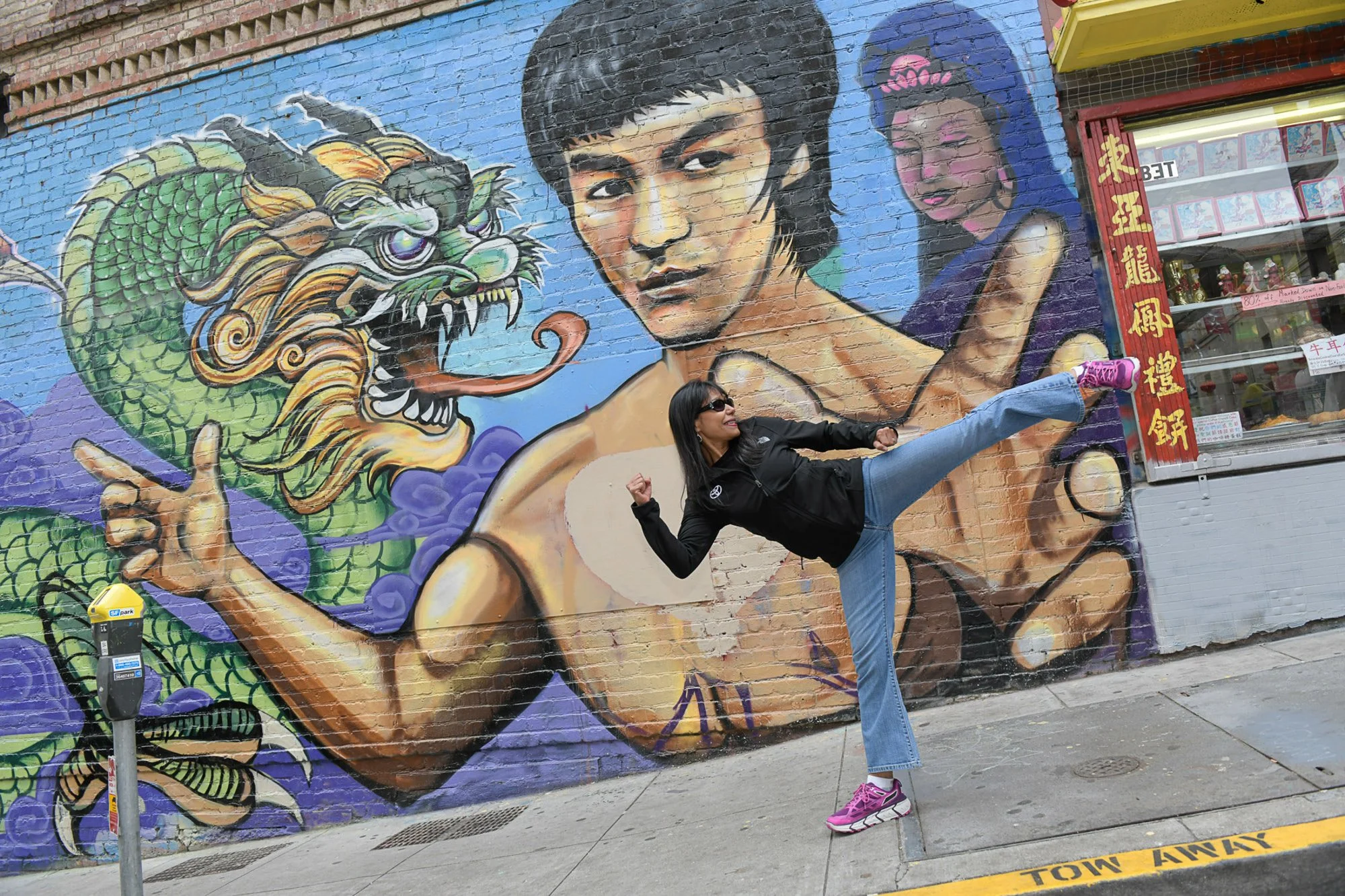 Woman in a black jacket, jeans, and pink sneakers mimicking a martial arts pose in front of a colorful mural on a brick wall, depicting a woman with short hair and a dragon.