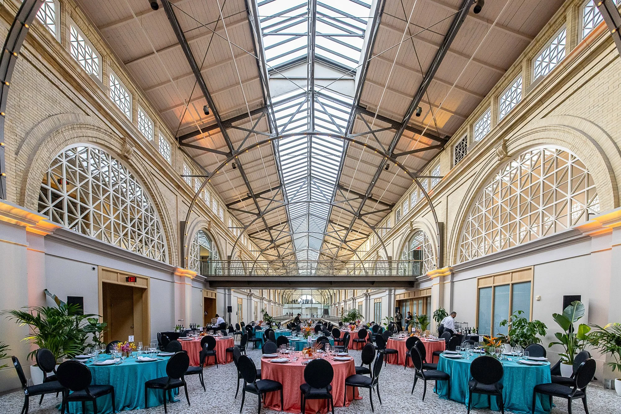 Interior of a large, elegant banquet hall with high arched ceilings, large windows, and a skylight, decorated for an event with round tables covered in colorful tablecloths and set with dinnerware, surrounded by black chairs, and green potted plants 