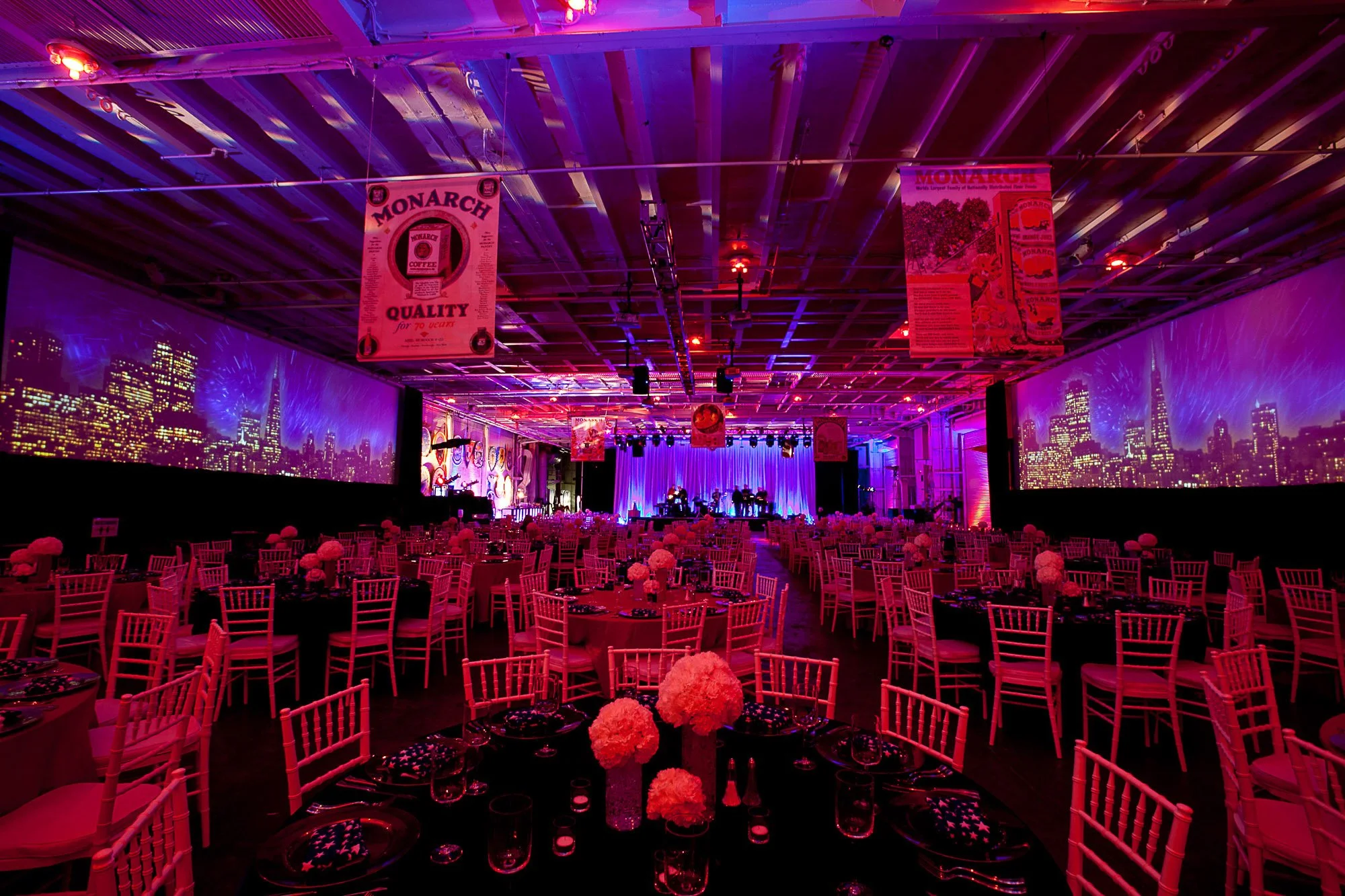 A large decorated banquet hall with tables and white chairs, pink and purple lighting, city skyline projections on the walls, and banners hanging from the ceiling.