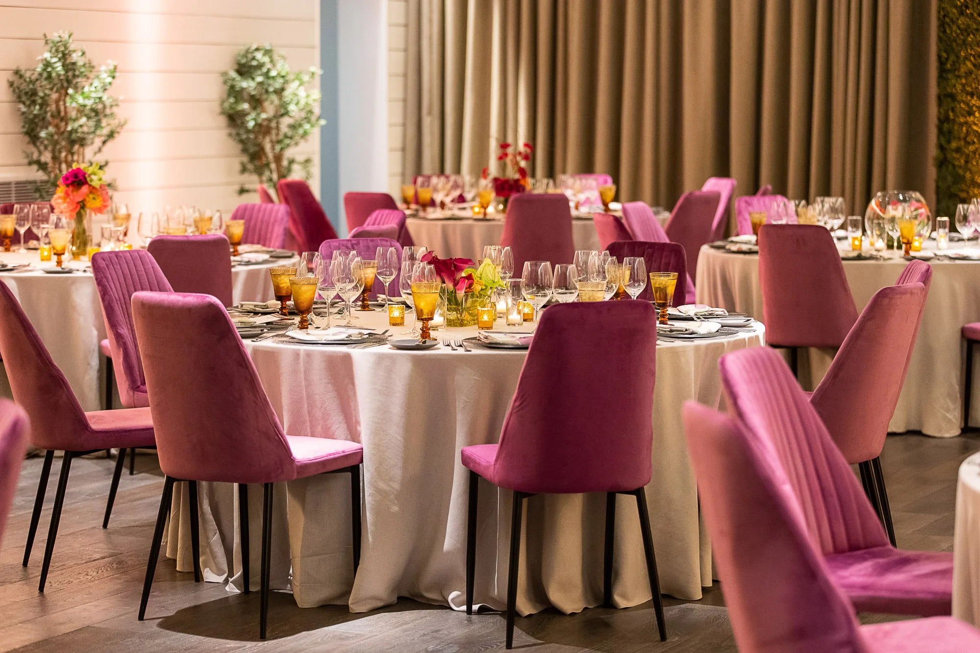 Elegant banquet hall with round tables covered in white tablecloths, set with wine glasses, candles, and floral centerpieces. Pink velvet chairs surround the tables, and beige curtains hang along the wall.