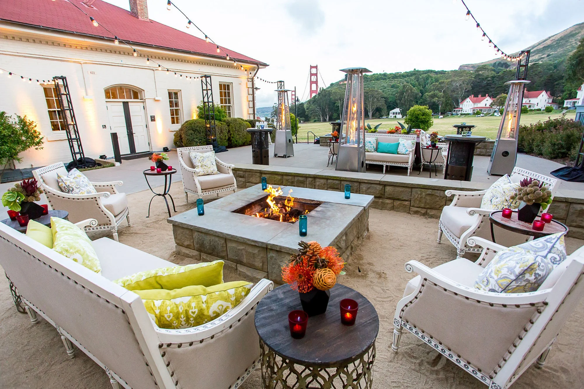 Outdoor patio with lounge chairs, a fire pit, string lights, and patio heaters. In the background, the Golden Gate Bridge and surrounding hills are visible.