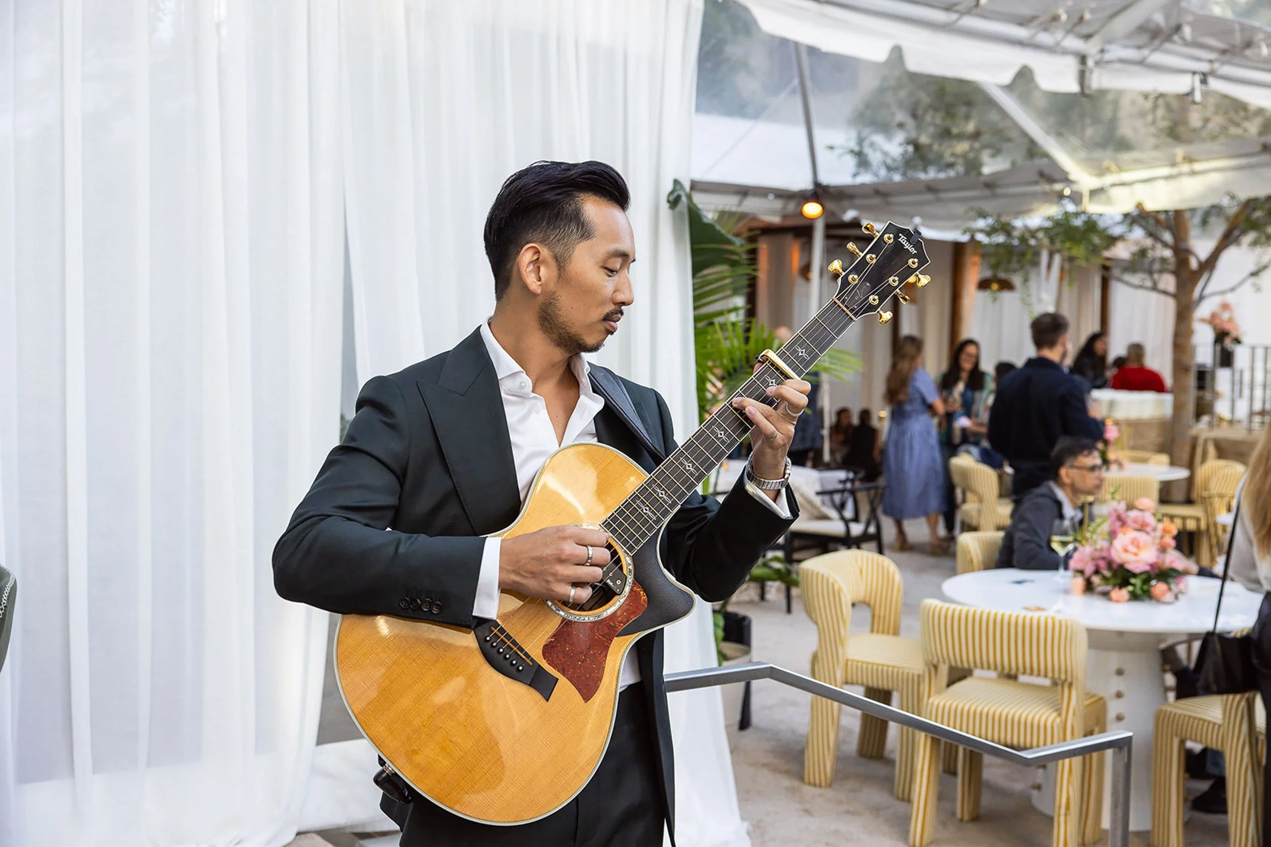 A man in a black suit playing an acoustic guitar indoors, with guests mingling in the background.