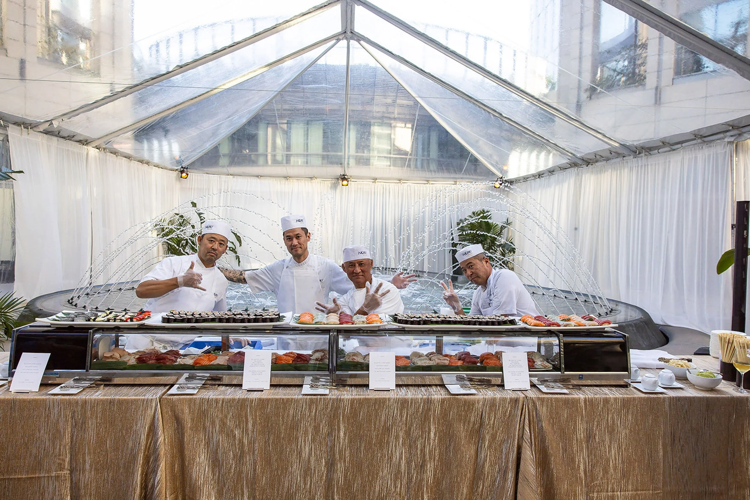 Four sushi chefs standing behind a sushi counter at a buffet, serving various sushi dishes, with a transparent tent canopy overhead and white curtains in the background.