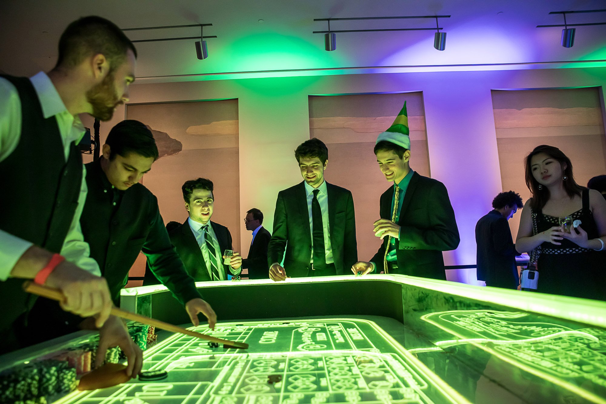 People dressed in formal attire playing on a glowing neon craps table at a casino-themed event, with colorful lighting and a woman wearing a party hat.