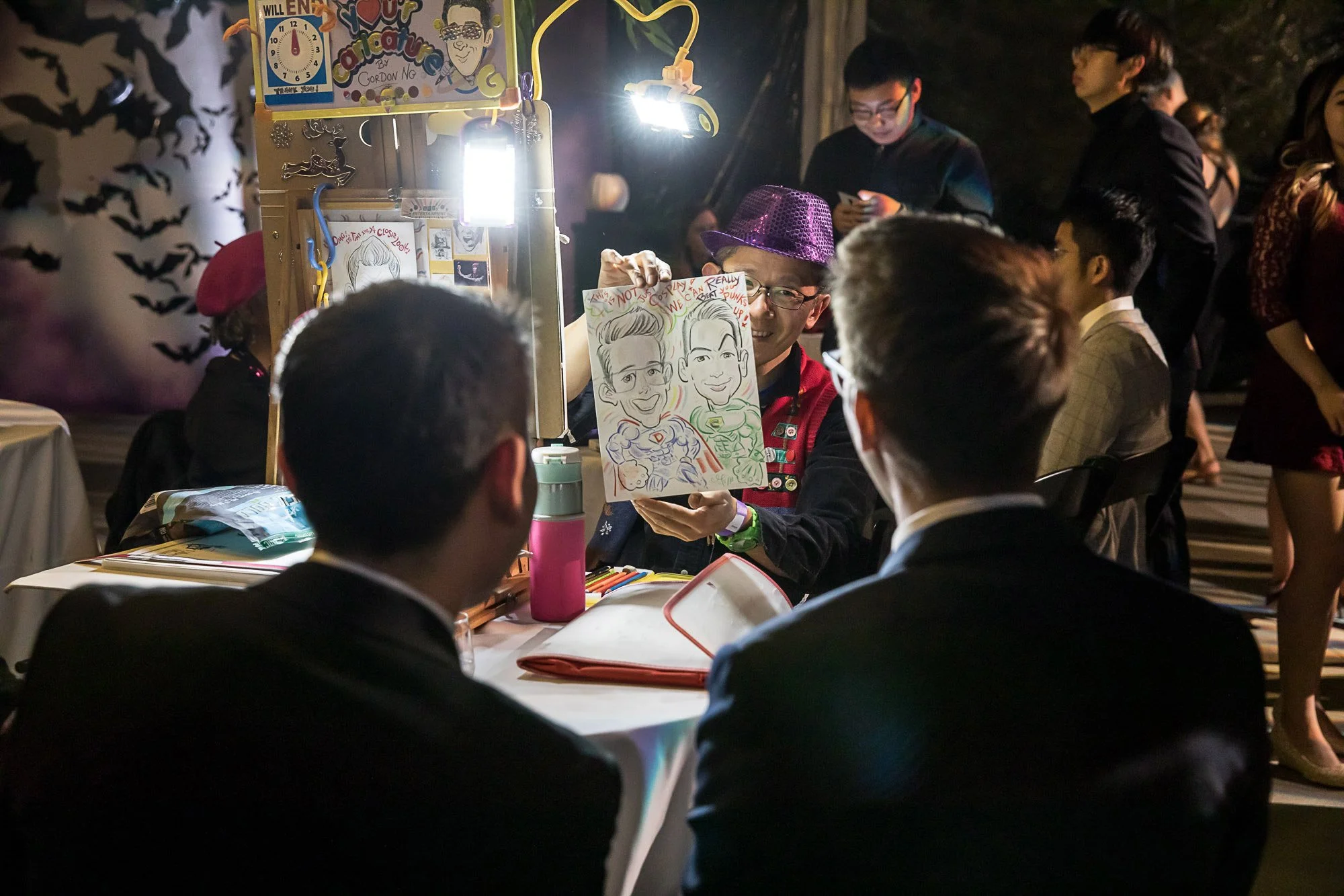 A man wearing a purple sequined hat and glasses is holding up a caricature portrait of himself and another person at a lively indoor event. Other people are gathered around, some looking at their phones. The caricature is drawn with vibrant colors an