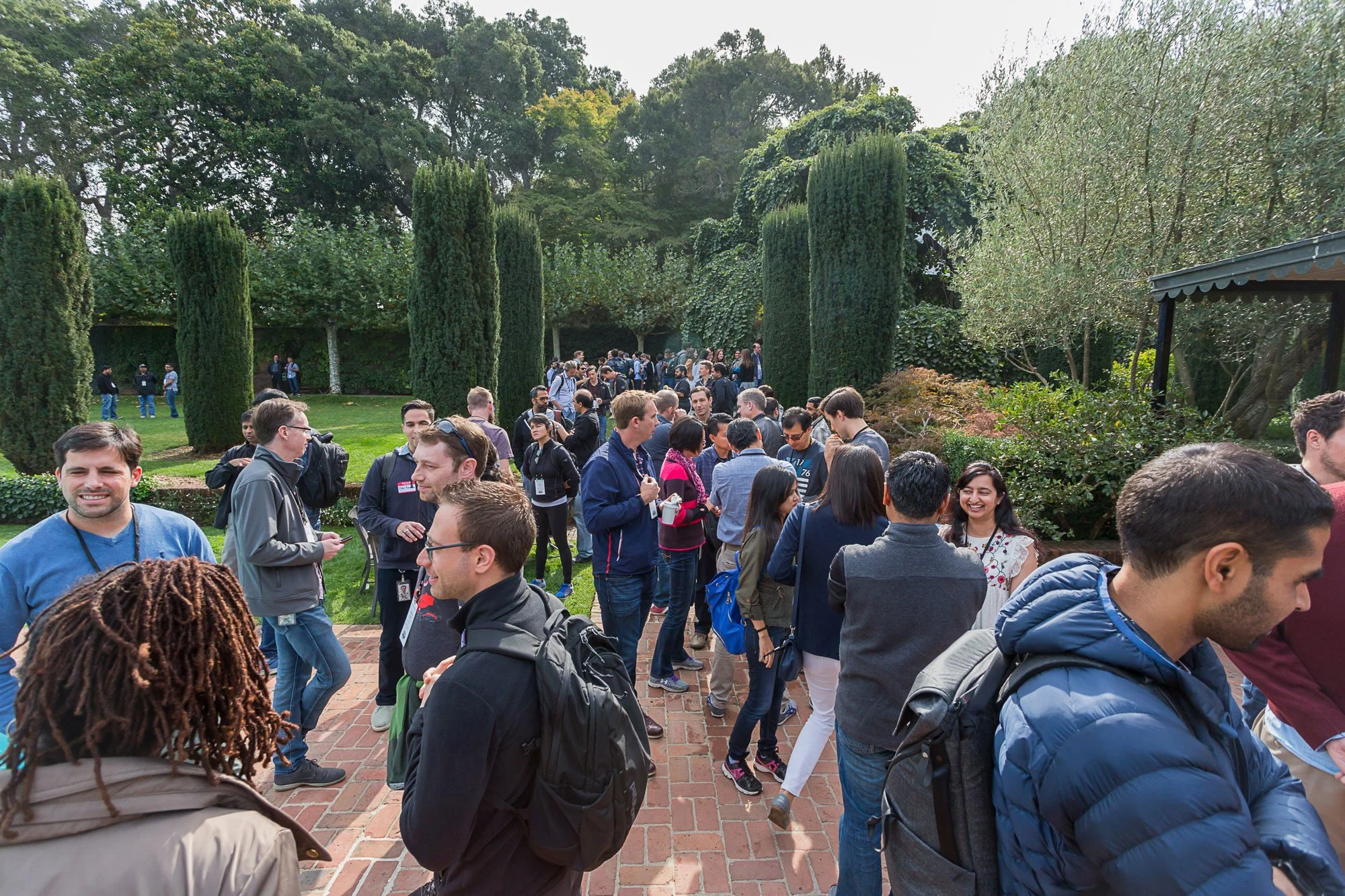 A group of people gathered outdoors in a park-like setting with tall trees and foliage, some standing in a queue or line, attending an event or gathering on a sunny day.