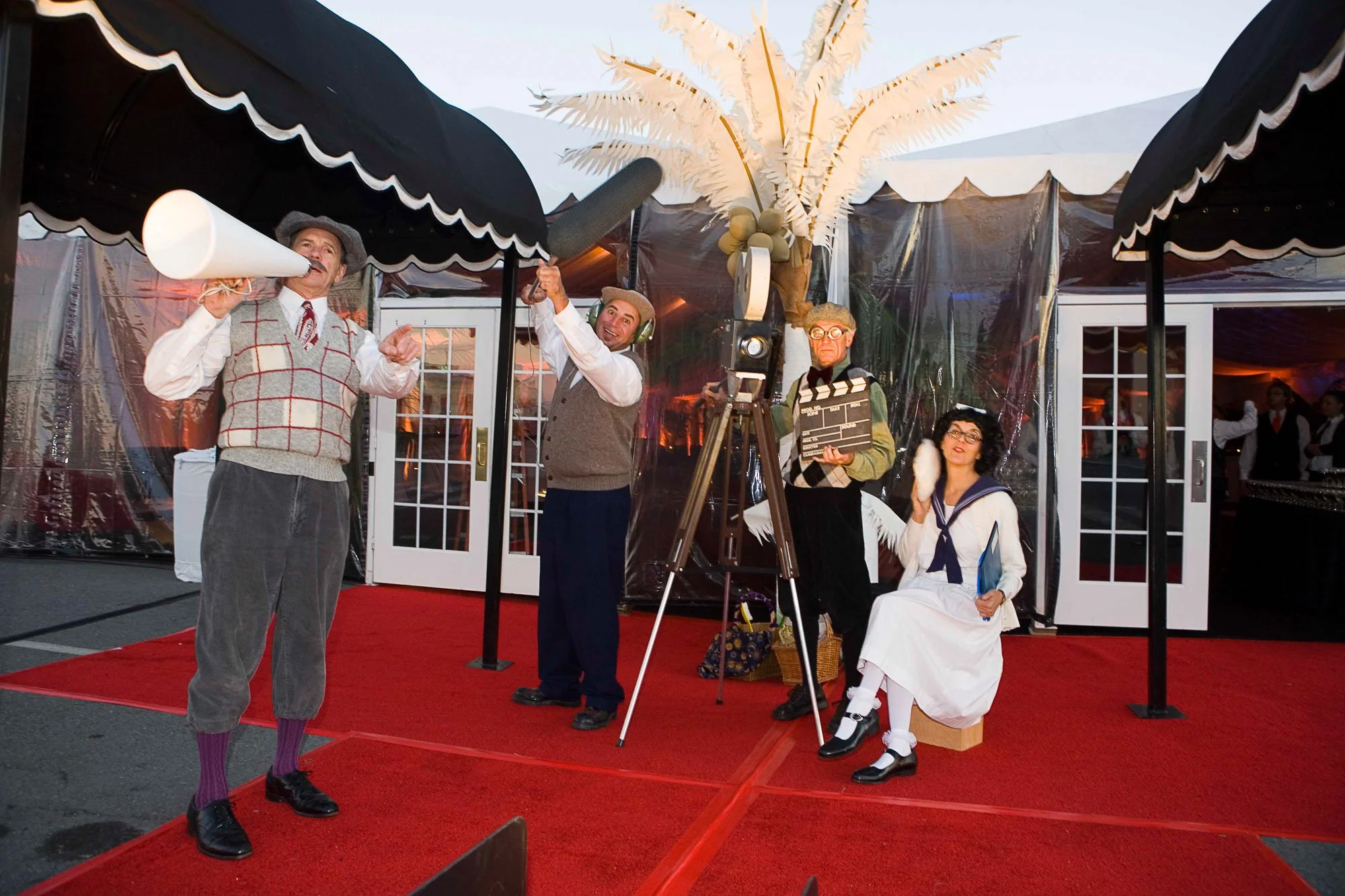 People dressed as vintage film characters on a red carpet with a backdrop of plastic-covered structures, including a palm tree prop and camera equipment, at a themed event.