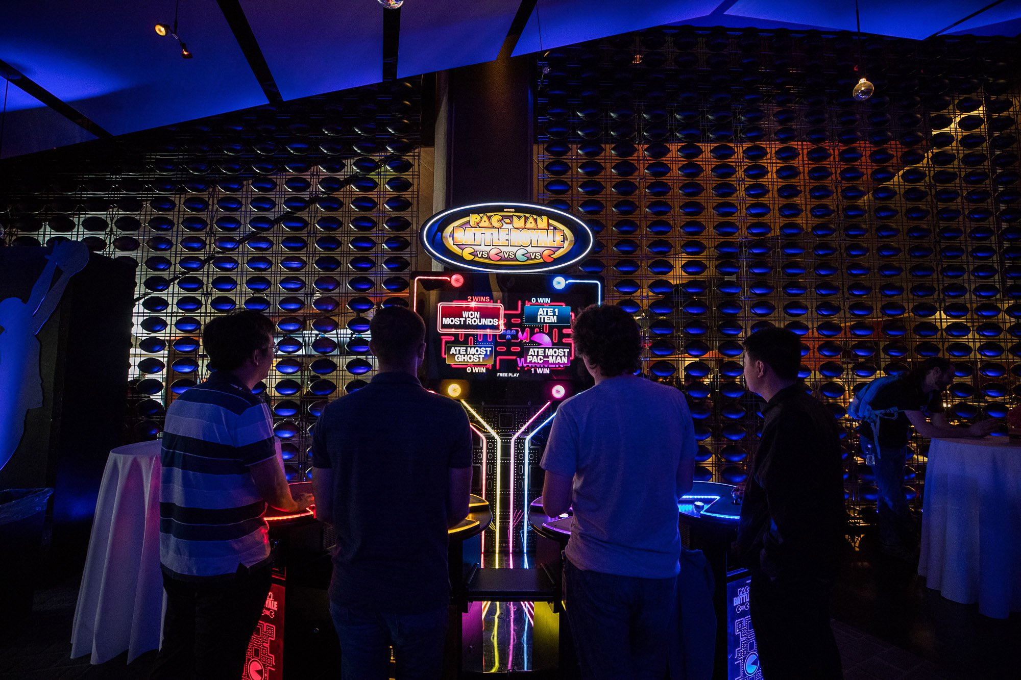 People playing Pac-Man Battle Royale arcade game at an event with neon lights and a decorative wall in the background.