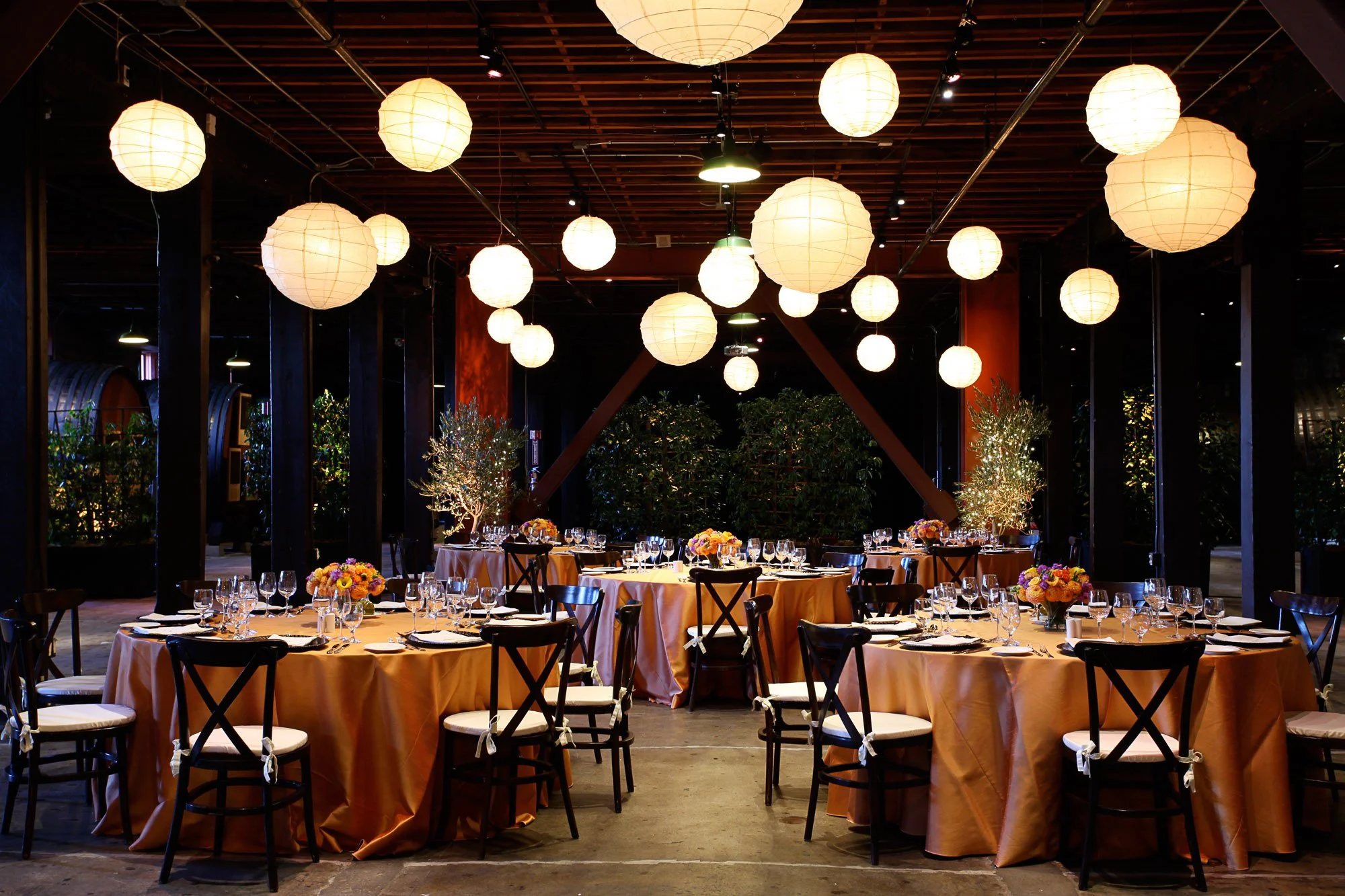 A decorated banquet hall with round tables covered in peach tablecloths, topped with floral centerpieces, neatly arranged glassware, and set silverware, under soft hanging lantern lights.