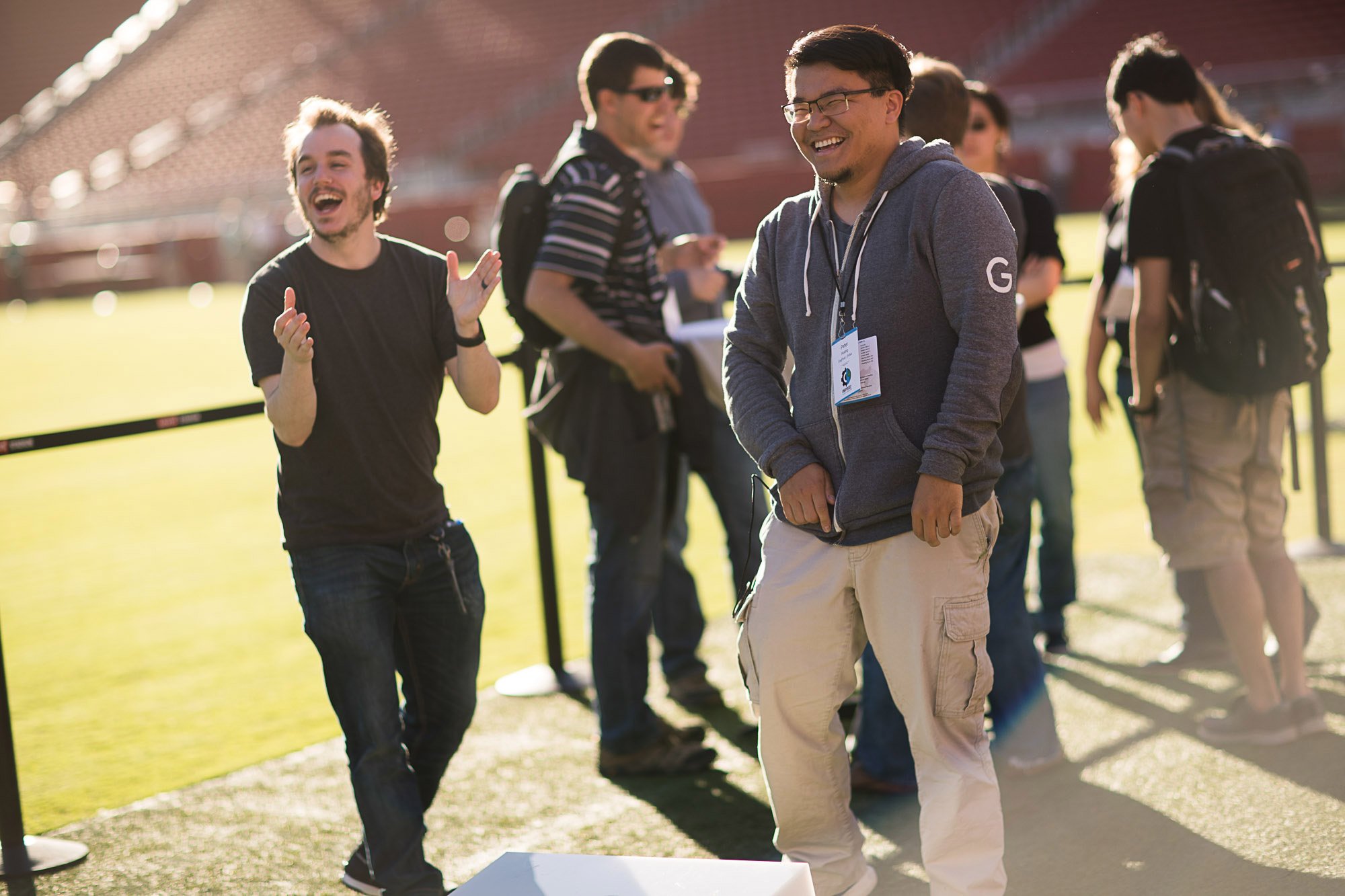 A group of young adults, mostly men, are standing outdoors near a baseball or football field, laughing and smiling. One man in the foreground with glasses, wearing a gray hoodie, is smiling with his eyes closed. Others are engaged in conversation and