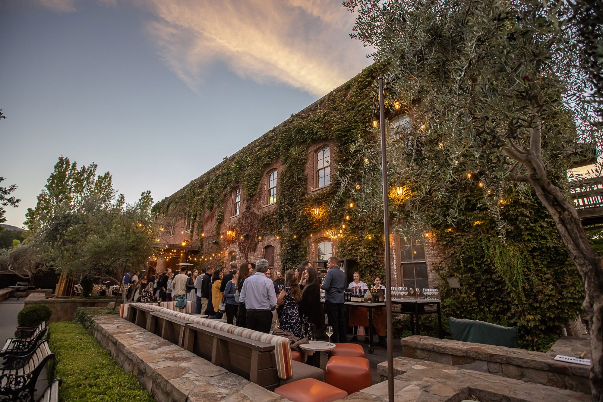 Outdoor social gathering at sunset with string lights and a moss-covered brick building in the background.