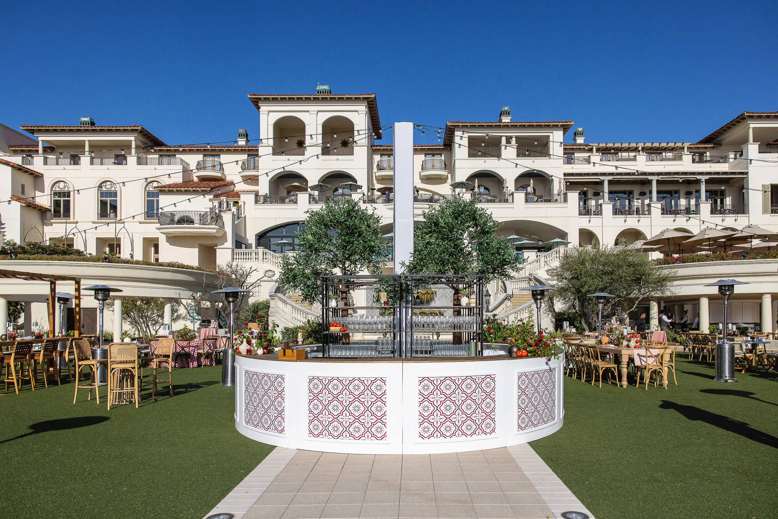 Outdoor banquet setup with tables and chairs, a central drink fountain, a multi-story building with balconies in the background, string lights, and clear blue sky.