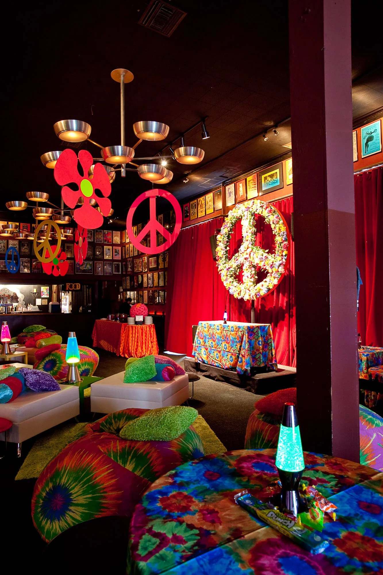 Colorful interior of a retro-themed room with tie-dye pillows, lava lamps, and psychedelic decor, featuring a stage with a peace sign and flower-shaped decorations.