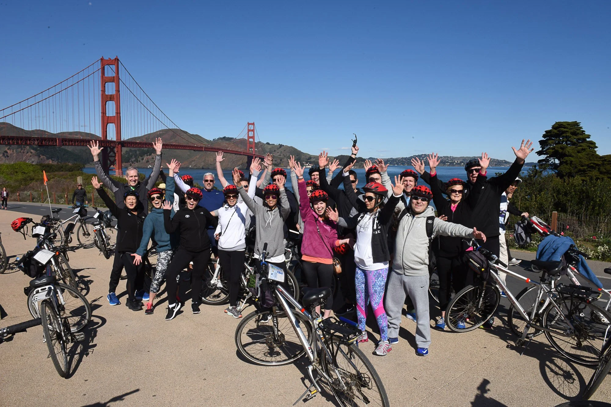Group of smiling people wearing cycling helmets and casual sports clothing, holding up their hands, standing next to bicycles in front of the Golden Gate Bridge on a sunny day.