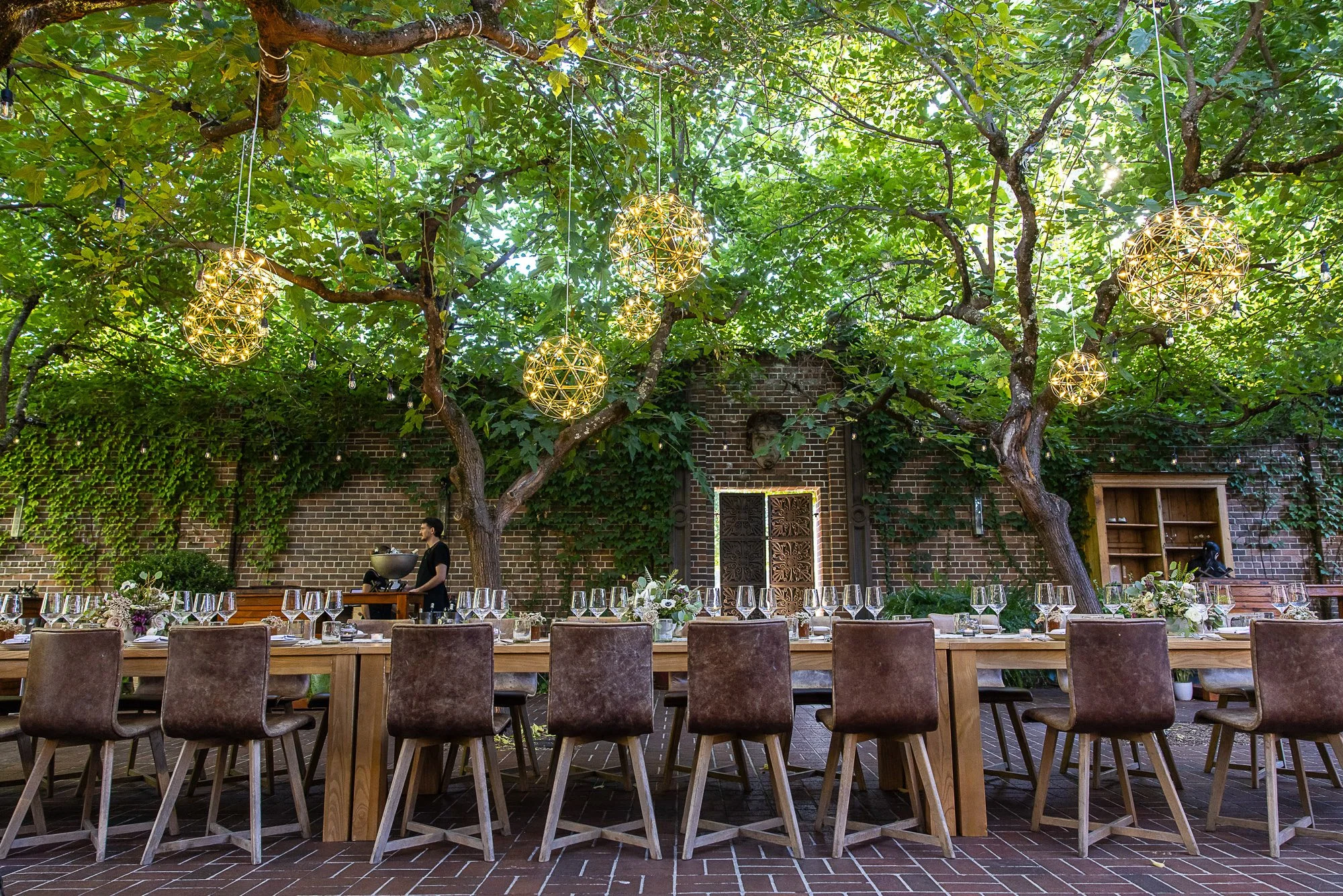 Outdoor dining setup under a tree with hanging spherical lights and string lights, brick wall background, setting for a formal event with tables, chairs, and floral centerpieces.
