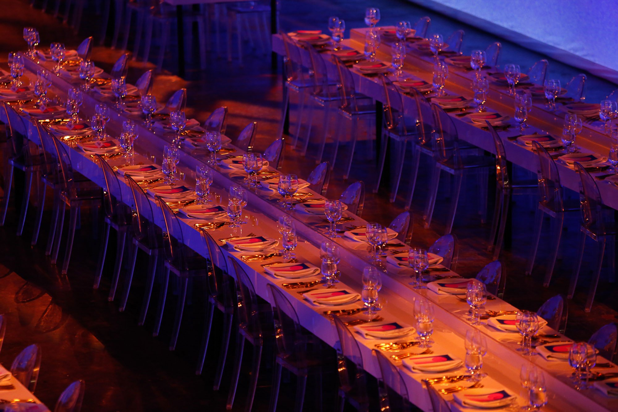 Long banquet table set for a formal event with plates, glasses, and cutlery, illuminated with warm, pink, and purple lighting, and surrounded by transparent chairs.