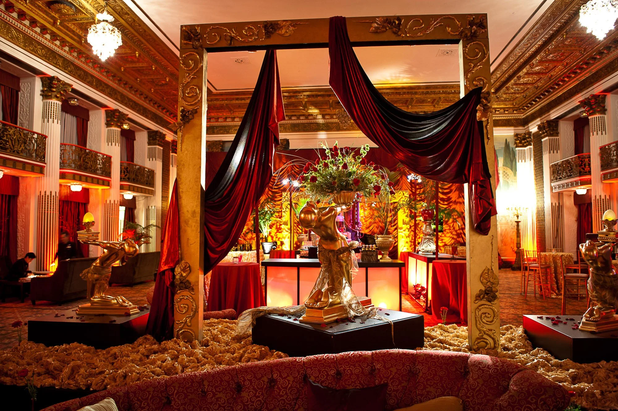 Elegant ballroom decorated with gold ornate details, red drapes, flowers, and lighting, featuring a display with golden sculptures and a floral arrangement in the center.