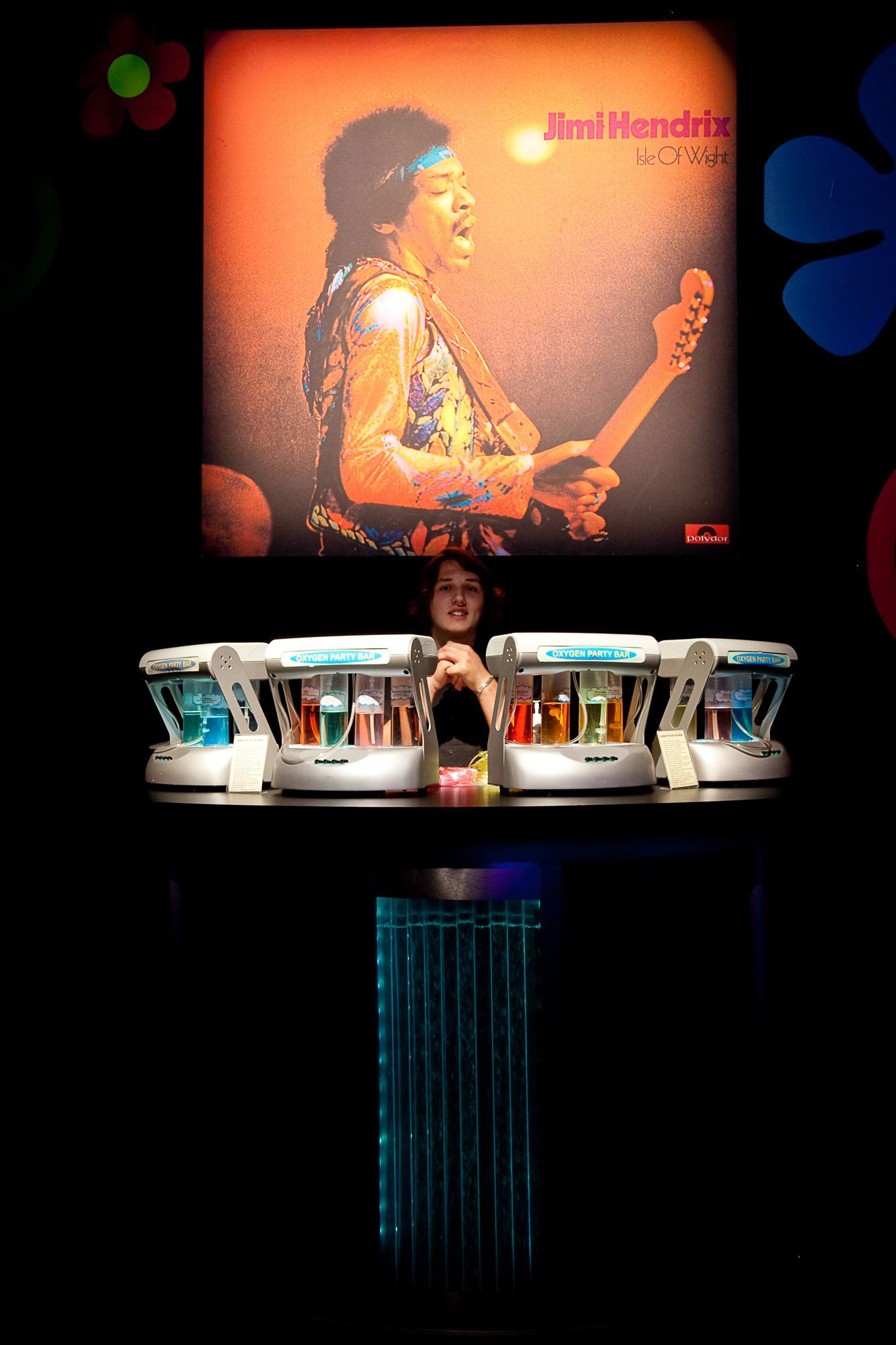 A woman standing behind a bar with colorful drink dispensers labeled 'Oxygen Party Bar' in a dark setting. Behind her is a large, projected image of Jimi Hendrix playing an electric guitar, with a vibrant background.
