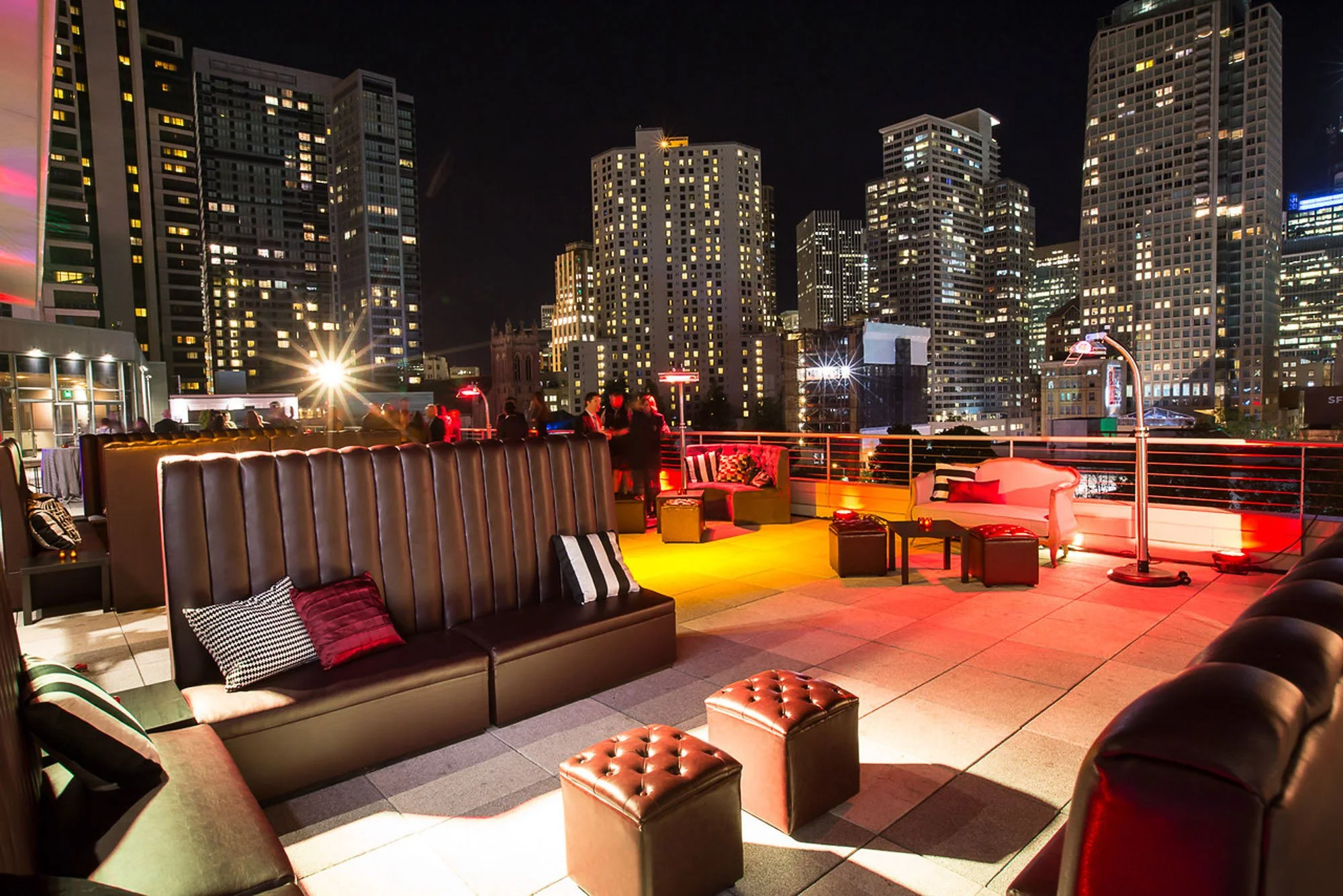 Rooftop lounge at night with city skyline in the background, featuring illuminated high-rise buildings and people socializing around tables with colorful lights and modern furniture.