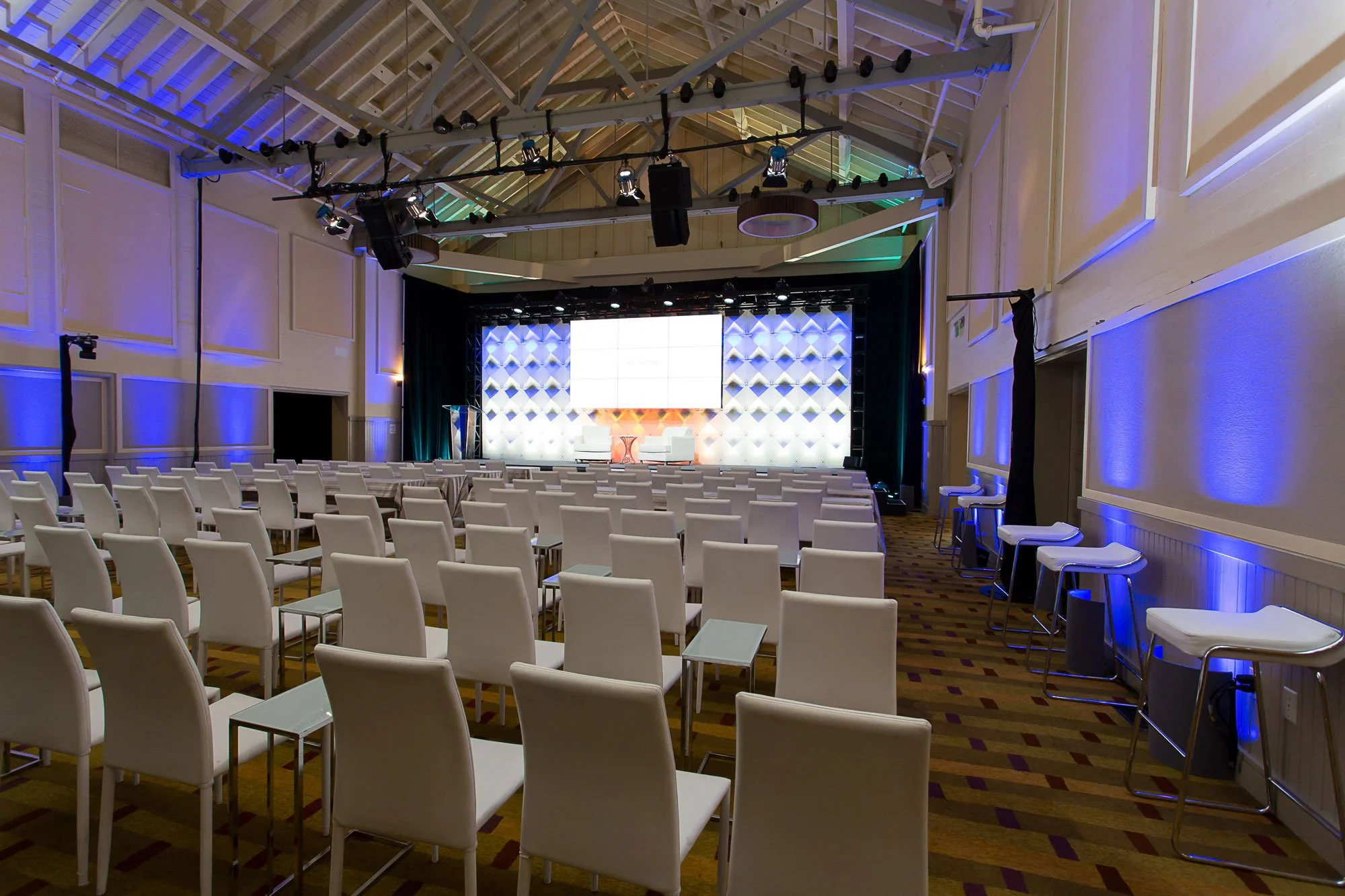 Empty conference or event hall with rows of white chairs facing a stage with a large white screen and decorative wall panels, painted in neutral tones with blue accent lighting.