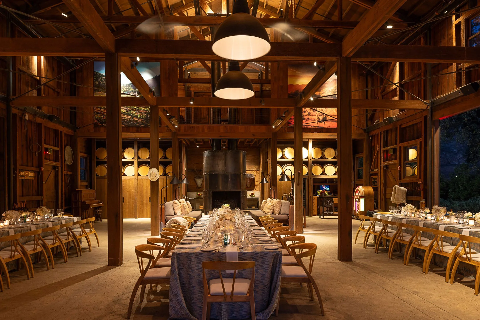 Interior of a rustic wooden banquet hall set for a dinner event with a long table, floral centerpieces, and dining chairs, illuminated by large hanging lamps in a barn-like structure.
